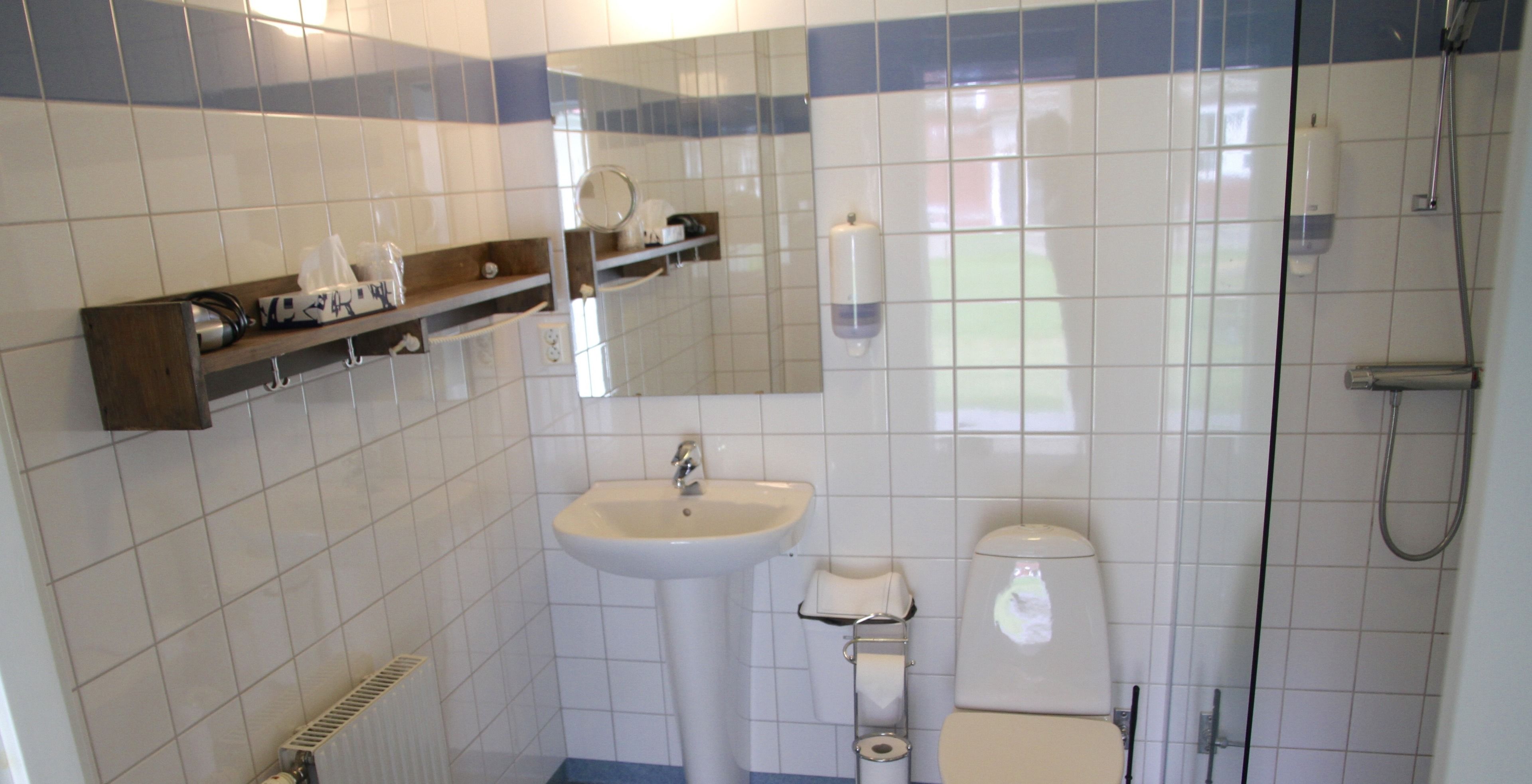 Un baño con lavabo, inodoro y ducha. Hay estantes con artículos de aseo y un espejo sobre el lavabo.