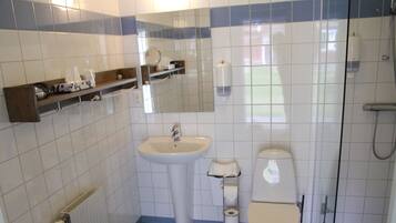 Un baño con lavabo, inodoro y ducha. Hay estantes con artículos de aseo y un espejo sobre el lavabo.