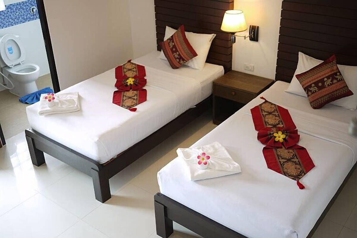 A hotel room with two single beds, each with white linens and red decorative pillows. There is a bedside table with a lamp, a small nightstand, and a view of a bathroom with a toilet and a blue tiled wall.