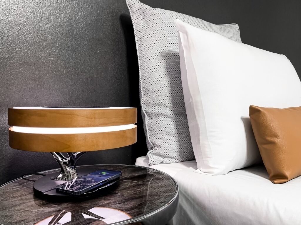 A modern lamp on a bedside table with a smartphone on it, a white pillow, and a grey pillow on a white sofa against a dark wall.