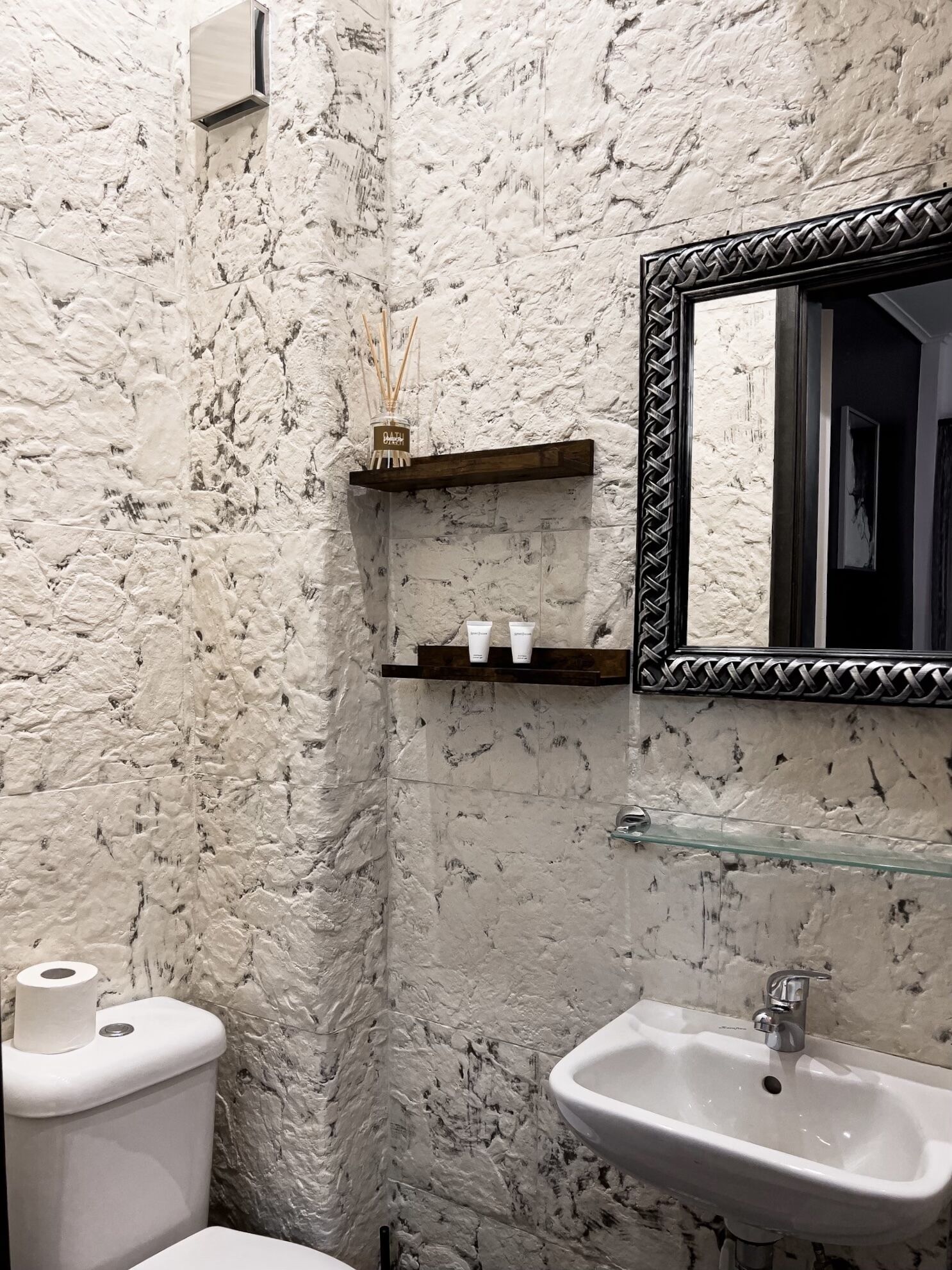 A bathroom with a marble wall, a toilet, a sink, a mirror, and a shelf with toiletries.