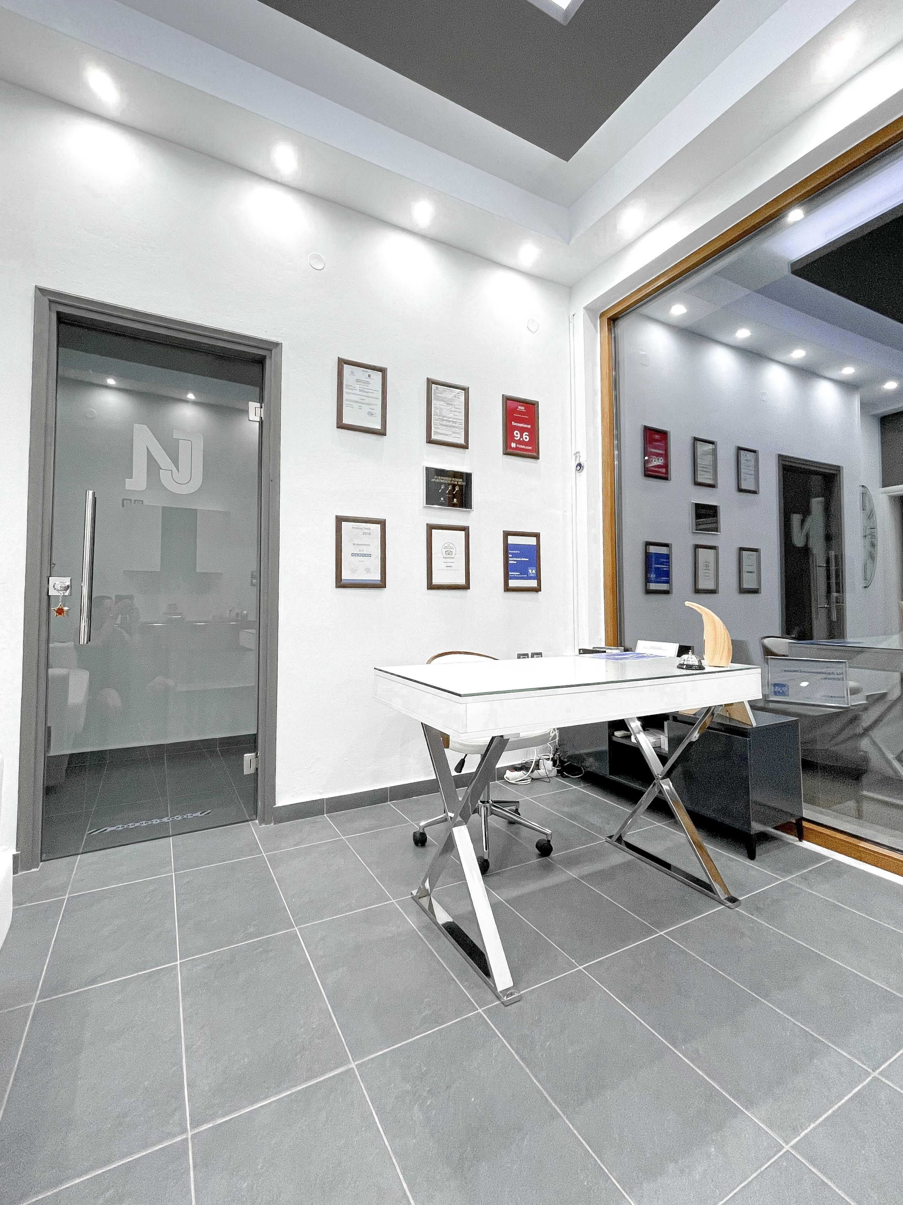 Un interior de oficina moderno con un escritorio de vidrio, certificados enmarcados en la pared y un espejo grande que refleja la habitación.