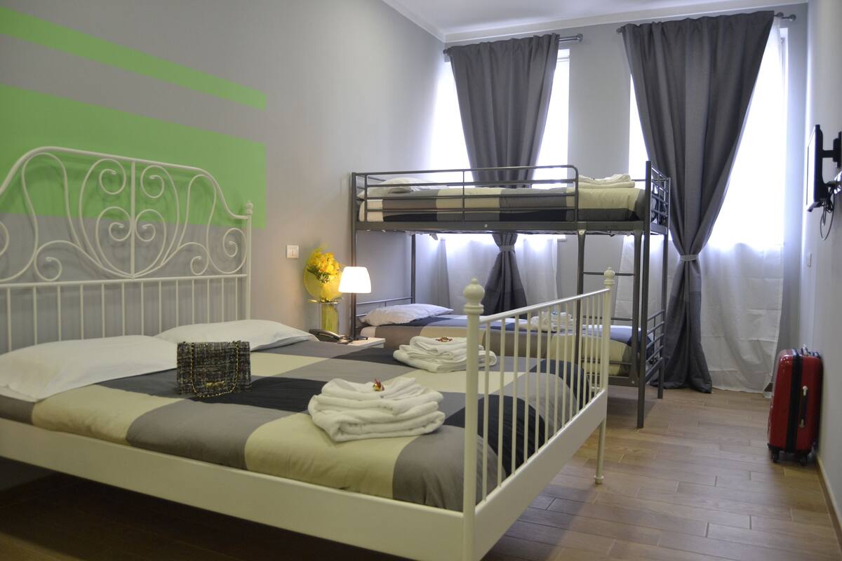 A hotel room with a bunk bed, a bedside table with a lamp, a suitcase, and a wall-mounted TV.