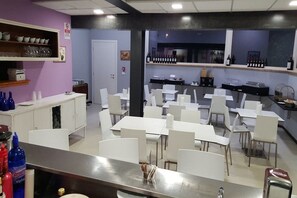 A restaurant with white tables and chairs, a bar area with bottles, and a shelf with cups and glasses.