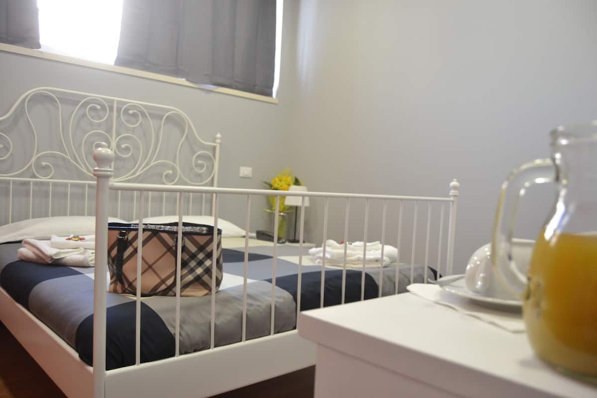 A neatly made bed with a metal headboard, a bedside table with a pitcher of water, and a vase with flowers on the nightstand.