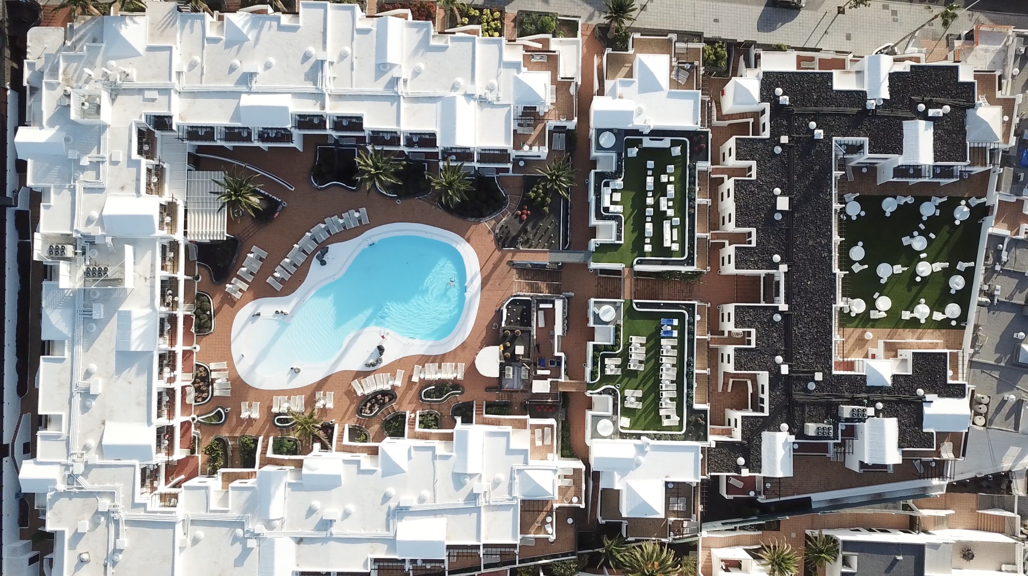 Vue aérienne d’un complexe hôtelier doté d’une piscine centrale, de plusieurs espaces de détente et d’un jardin bien entretenu.
