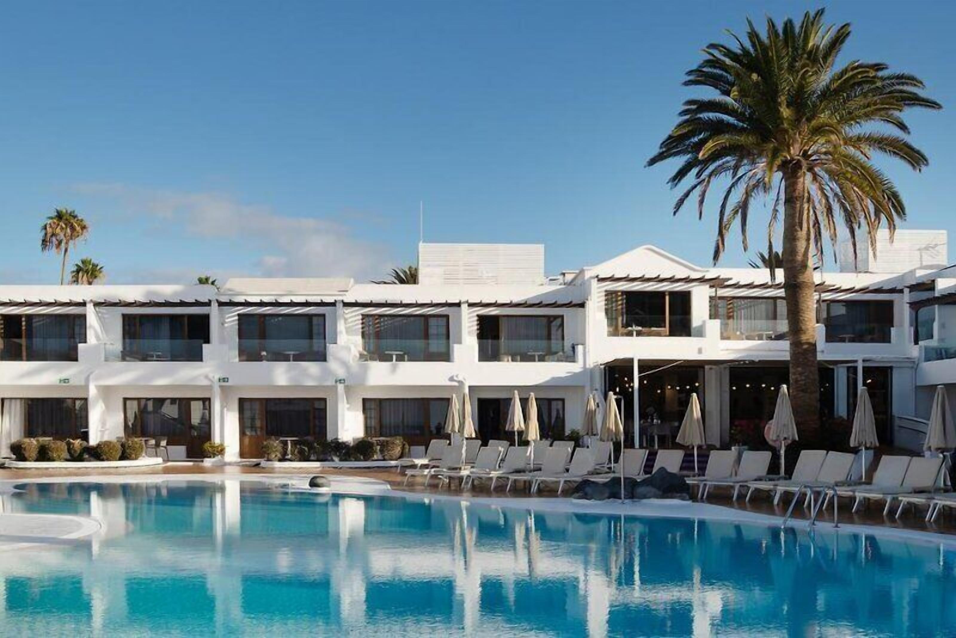 Un albergo con piscina, lettini, ombrelloni e palme.