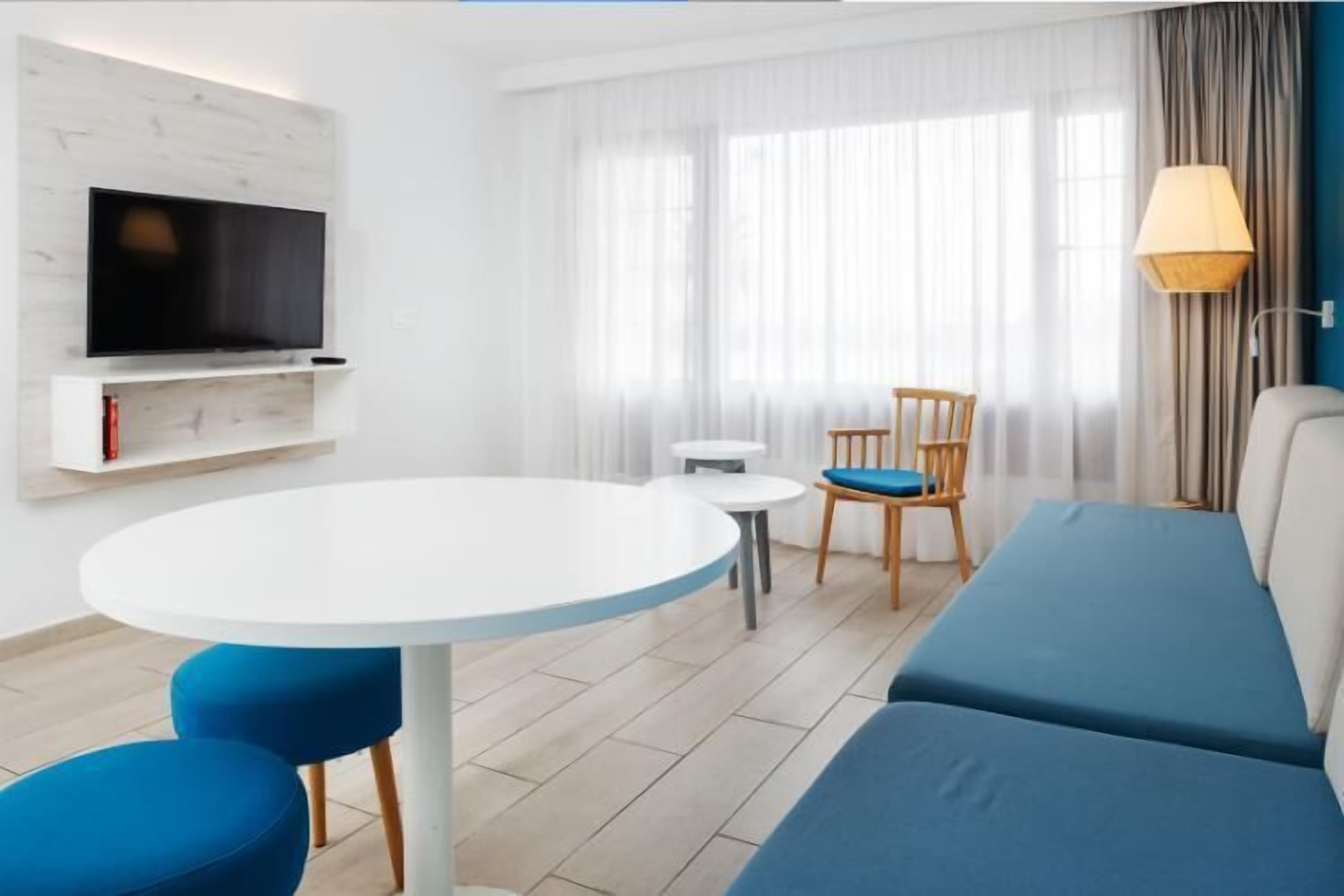 A modern living room with a flat-screen TV, a round white table, blue upholstered chairs, a sofa, and a lamp.