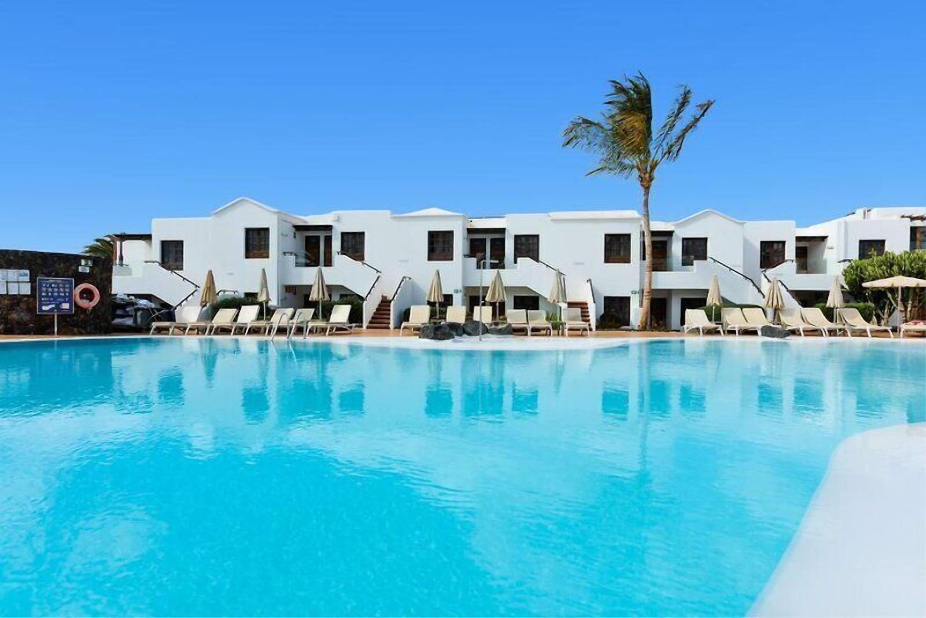 A poolside view of a hotel with white buildings, sun loungers, and a palm tree.