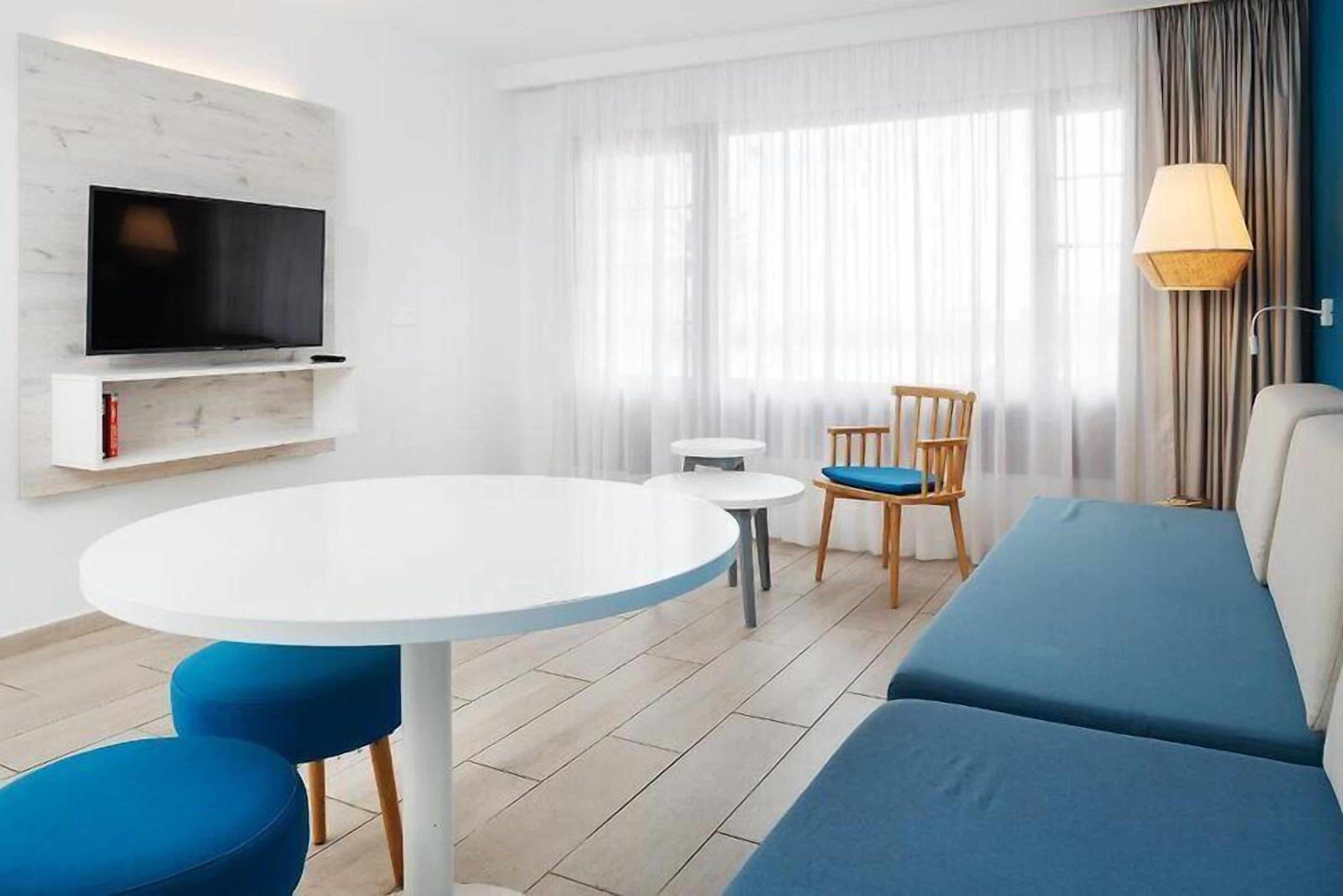 Ett modernt hotellrum med en platt-TV som är monterad på en vit vägg, ett runt matbord med blå stolar, en blå soffa och ett litet vitt bord.