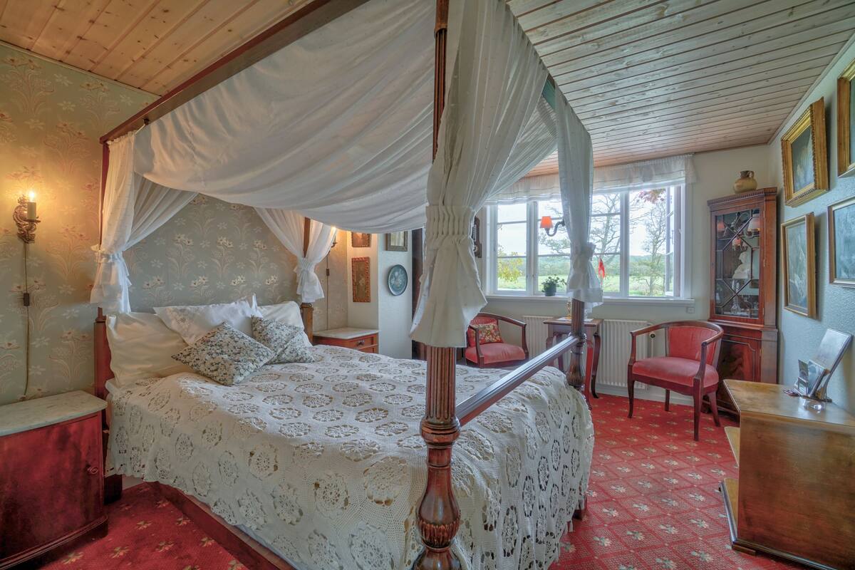 A four-poster canopy bed with a lace bedspread, a wooden nightstand, a red chair, and a wooden dresser with a mirror.