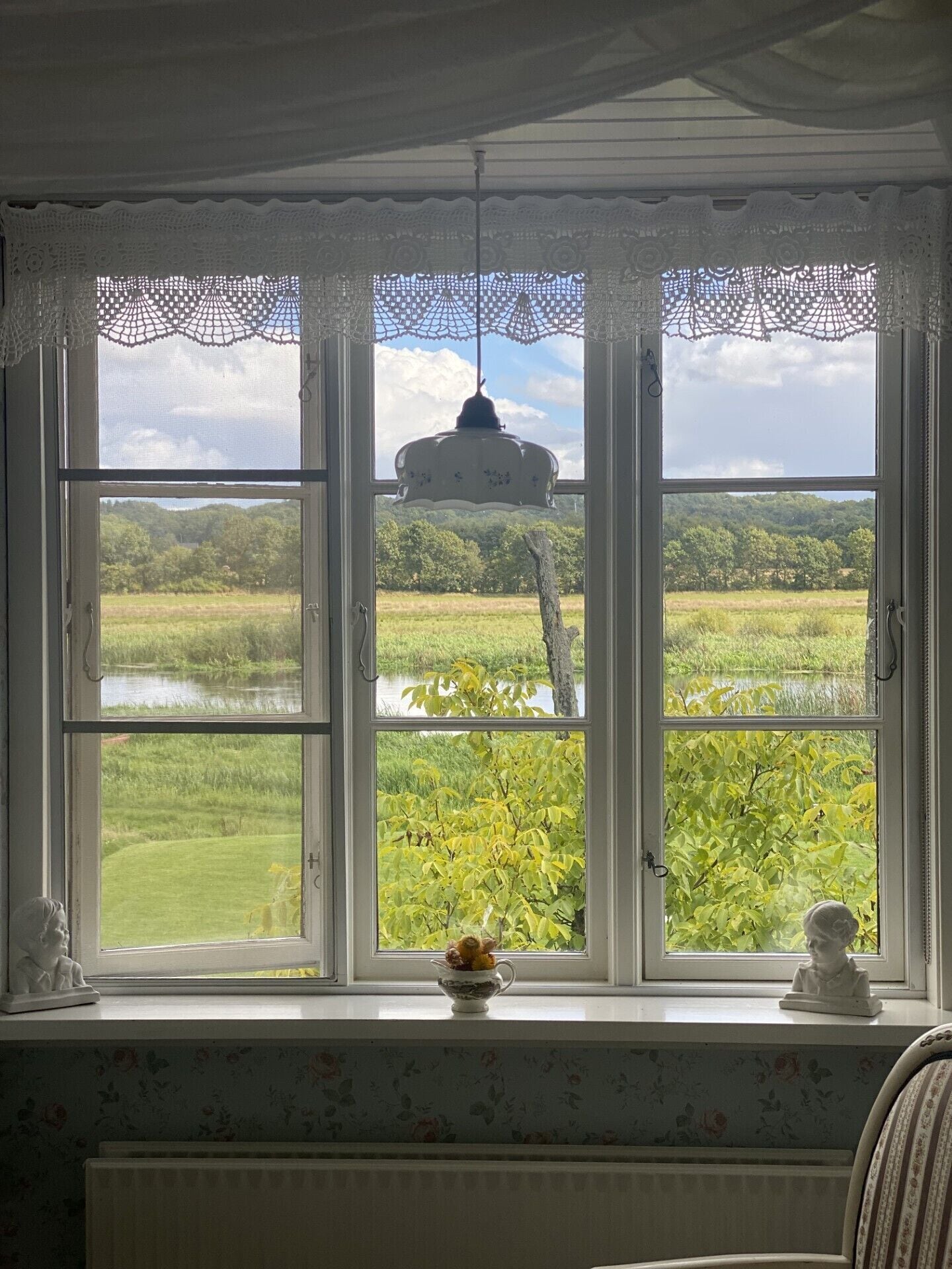Una habitación con un gran ventanal que da a un paisaje verde, una lámpara colgante y un pequeño florero con flores en el alféizar.