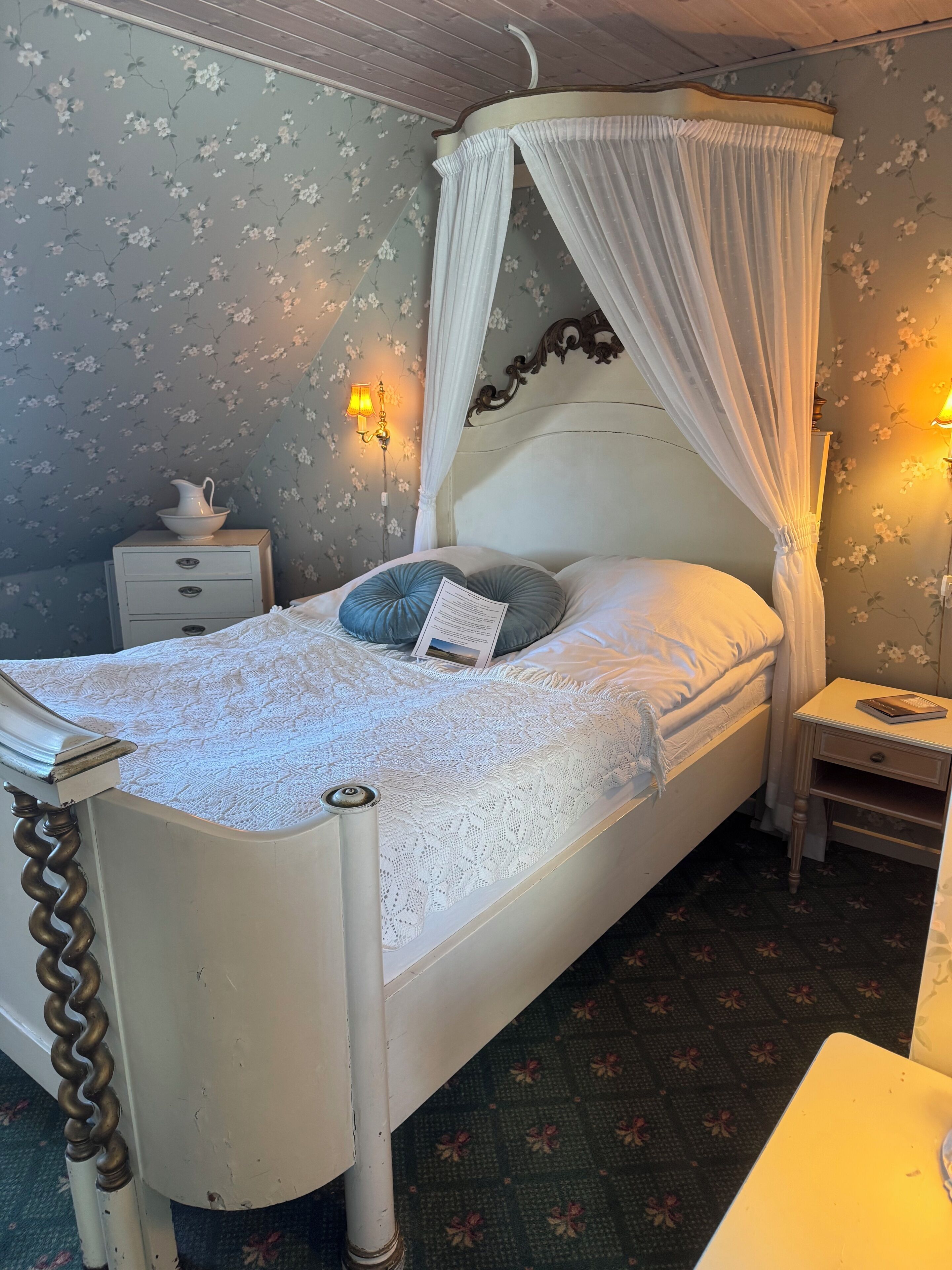 Una cama con dosel, una mesita de noche con lámpara, una mesita auxiliar y una pared con papel tapiz floral.
