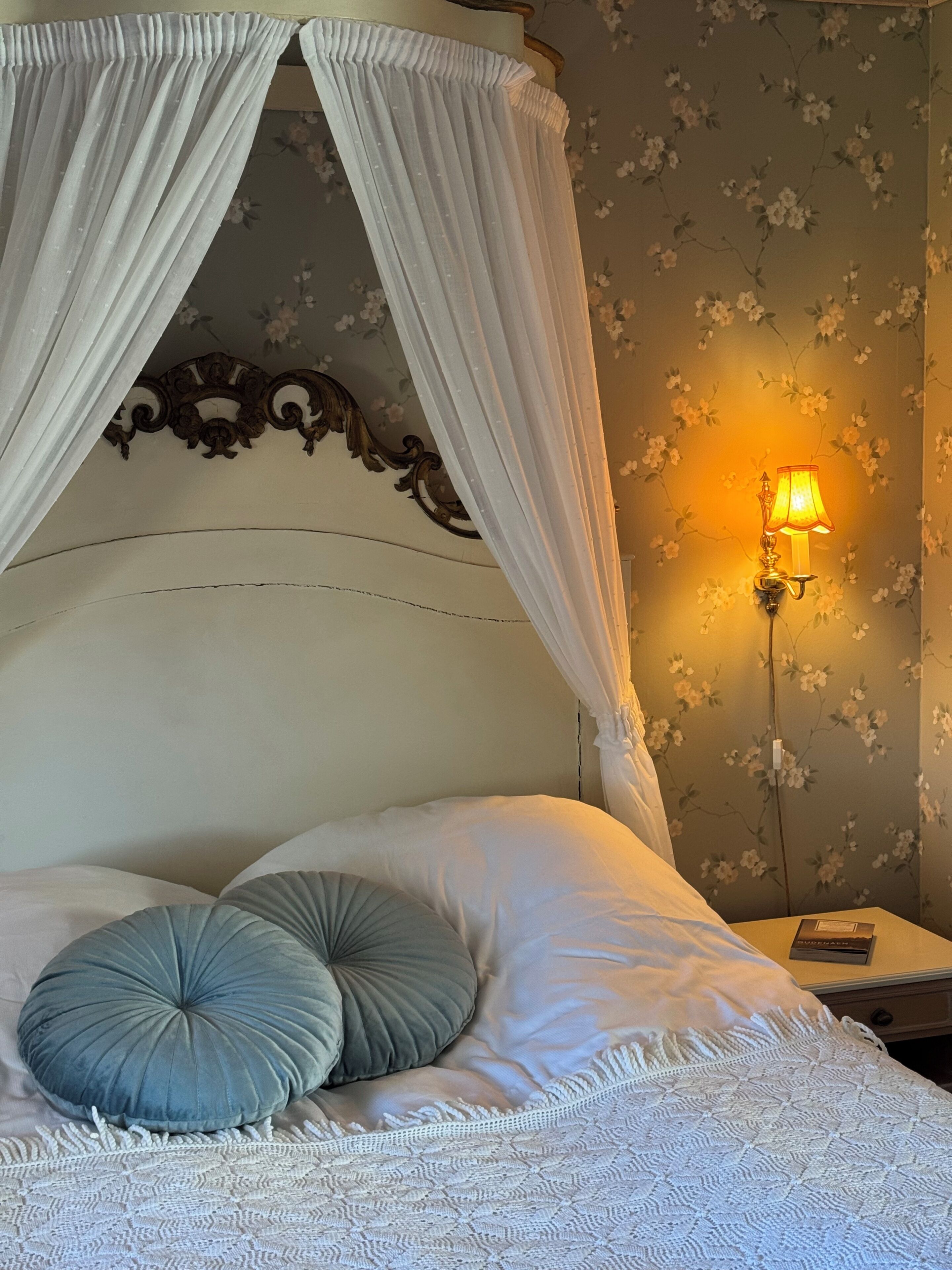 Un dormitorio con cama con dosel, papel tapiz floral, una mesita de noche con lámpara y un libro.