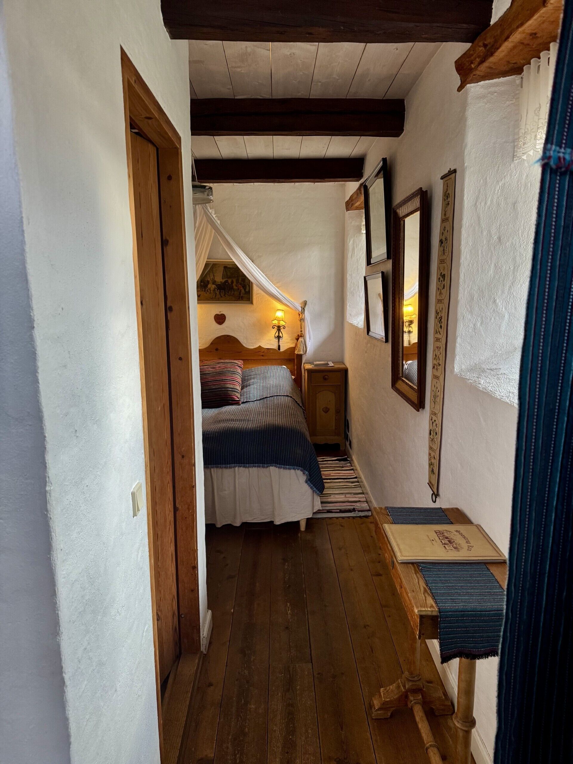Un pasillo estrecho que conduce a un dormitorio con una cama, una mesita de noche, una lámpara y un banco de madera.
