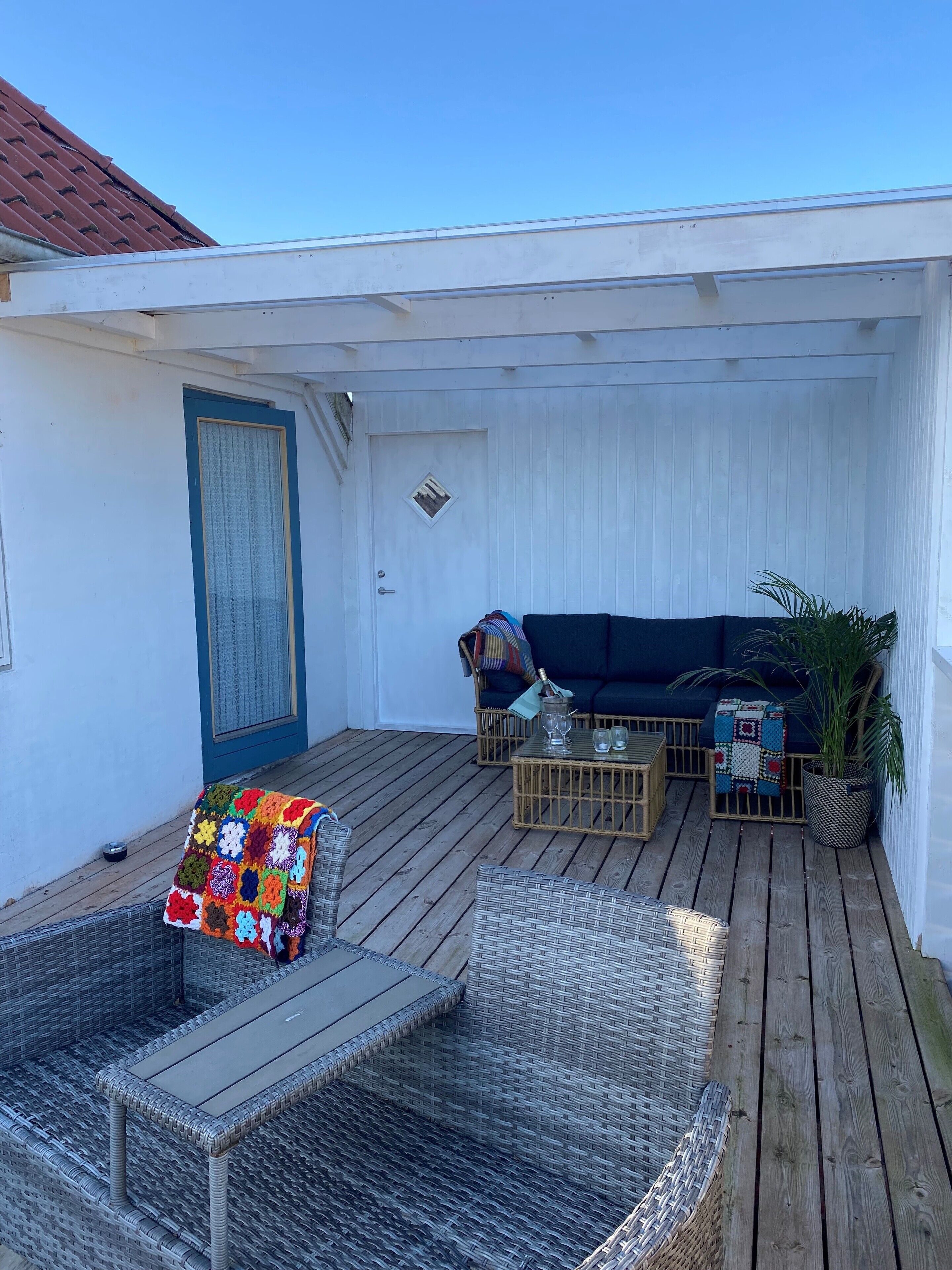 En overdækket udendørs siddeplads med et flettet bord, en polstret bænk og en mørk sofa.