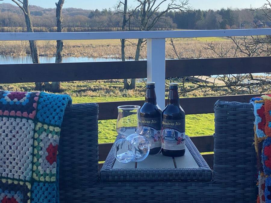 Una mesa de mimbre con dos botellas de Rosstrap Ale, un vaso y una manta colorida de ganchillo, sobre una terraza con vistas a un lago.