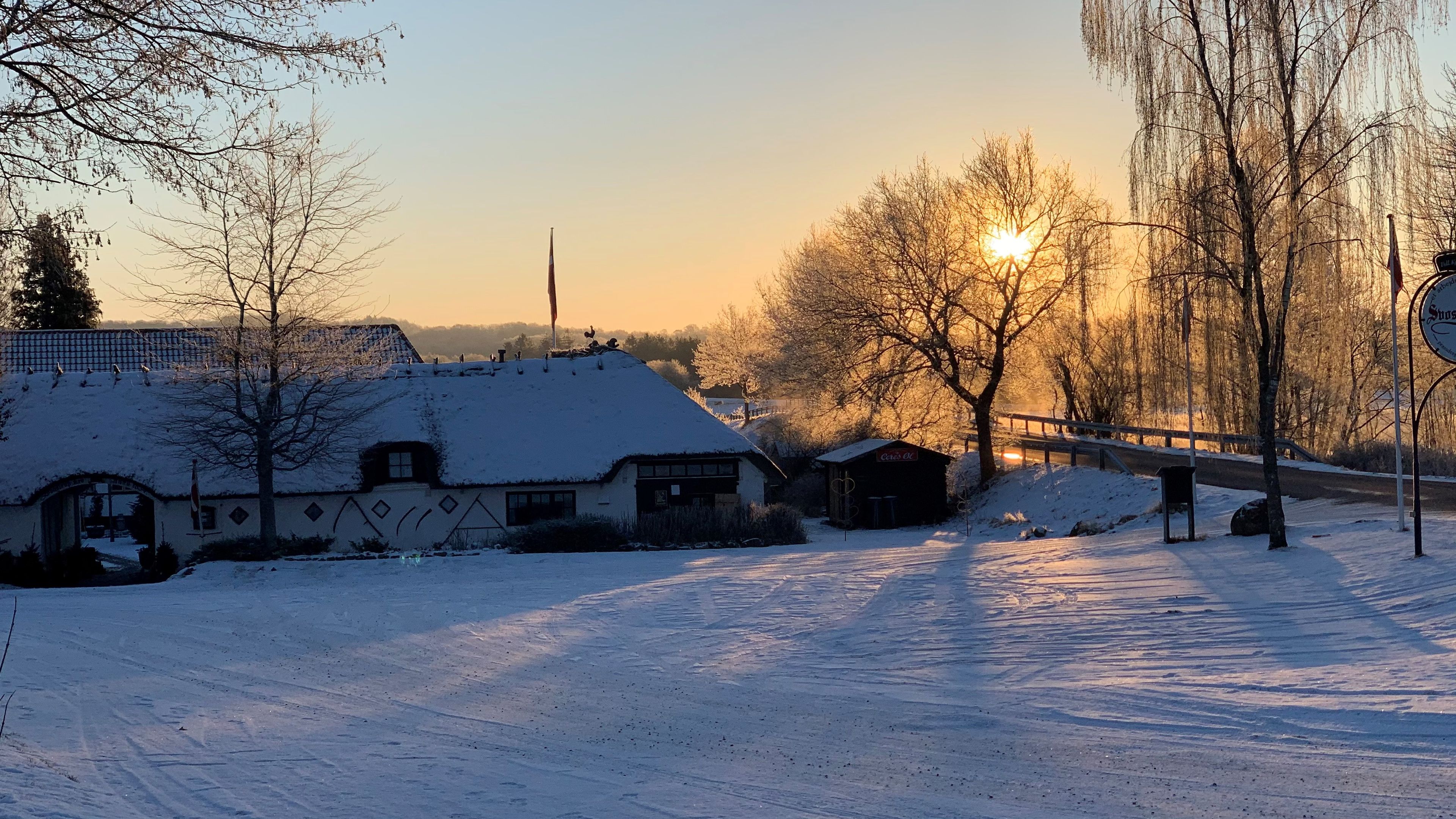 Lumipeitteinen maisema, jossa on perinteinen rakennus, puinen silta ja kirkas taivas.