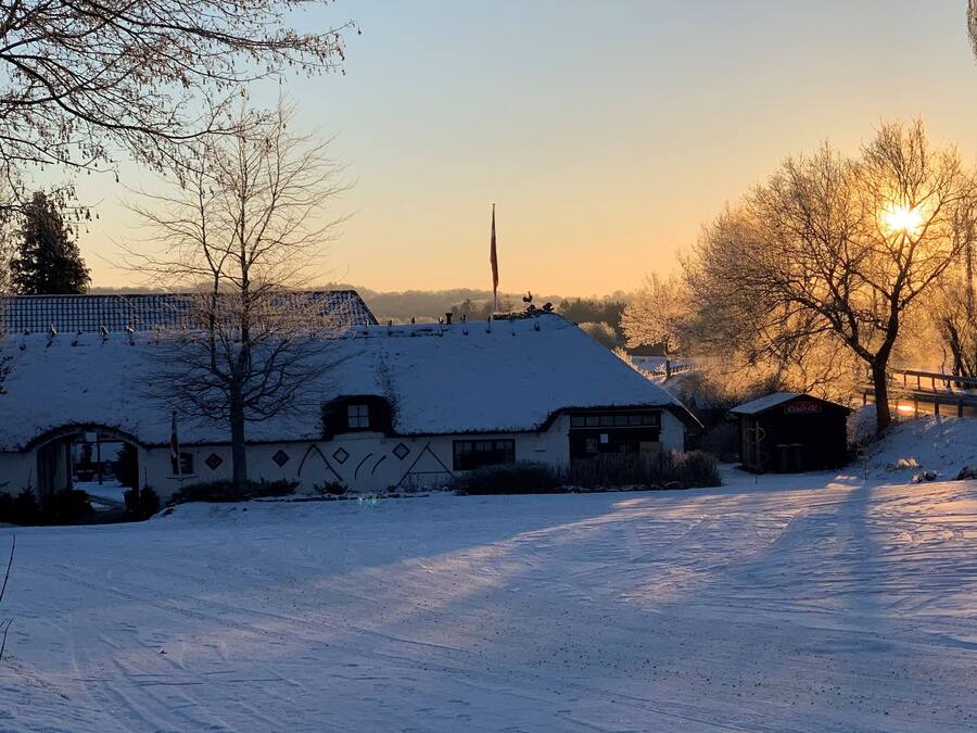 Lumipeitteinen maisema, jossa on perinteinen rakennus, puinen silta ja kirkas taivas.