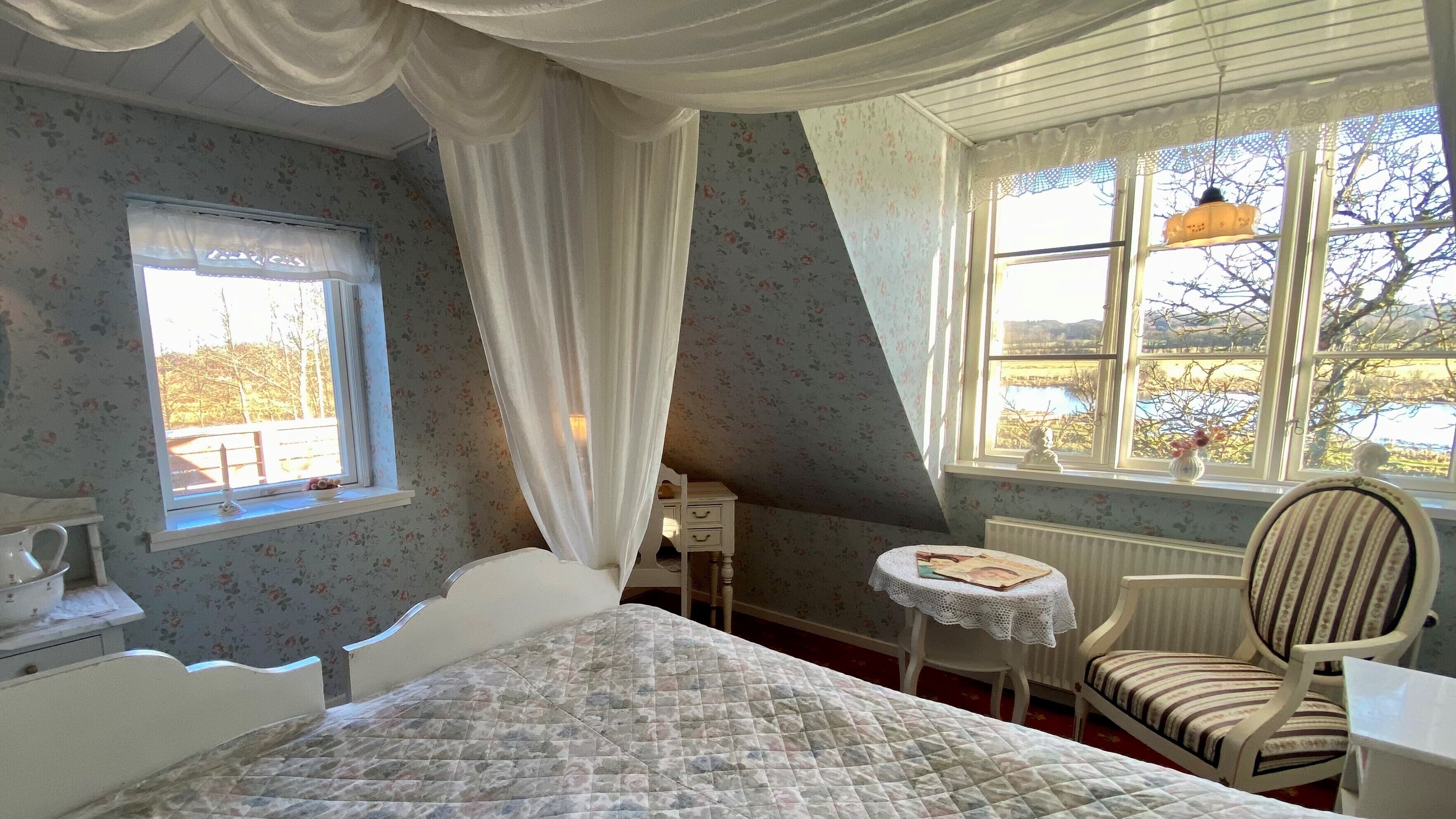 A bedroom with a bed, a chair, a small table, a window with curtains, and a view of the outdoors.