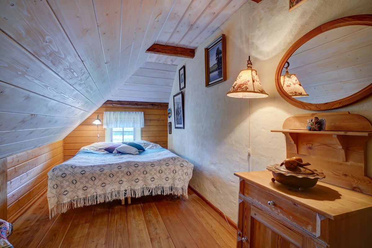 A cozy attic bedroom with a wooden sloped ceiling, a bed with a patterned bedspread, two wall-mounted lamps, and a wooden dresser with a mirror.