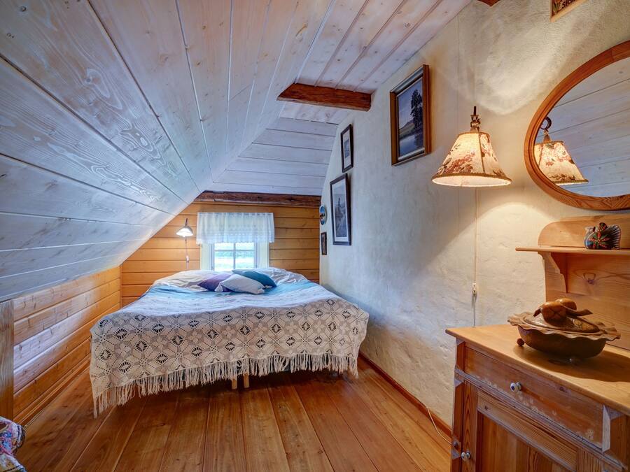 Un dormitorio acogedor en el ático con techo inclinado de madera, una cama con colcha estampada, dos lámparas fijadas a la pared y un aparador de madera con espejo.