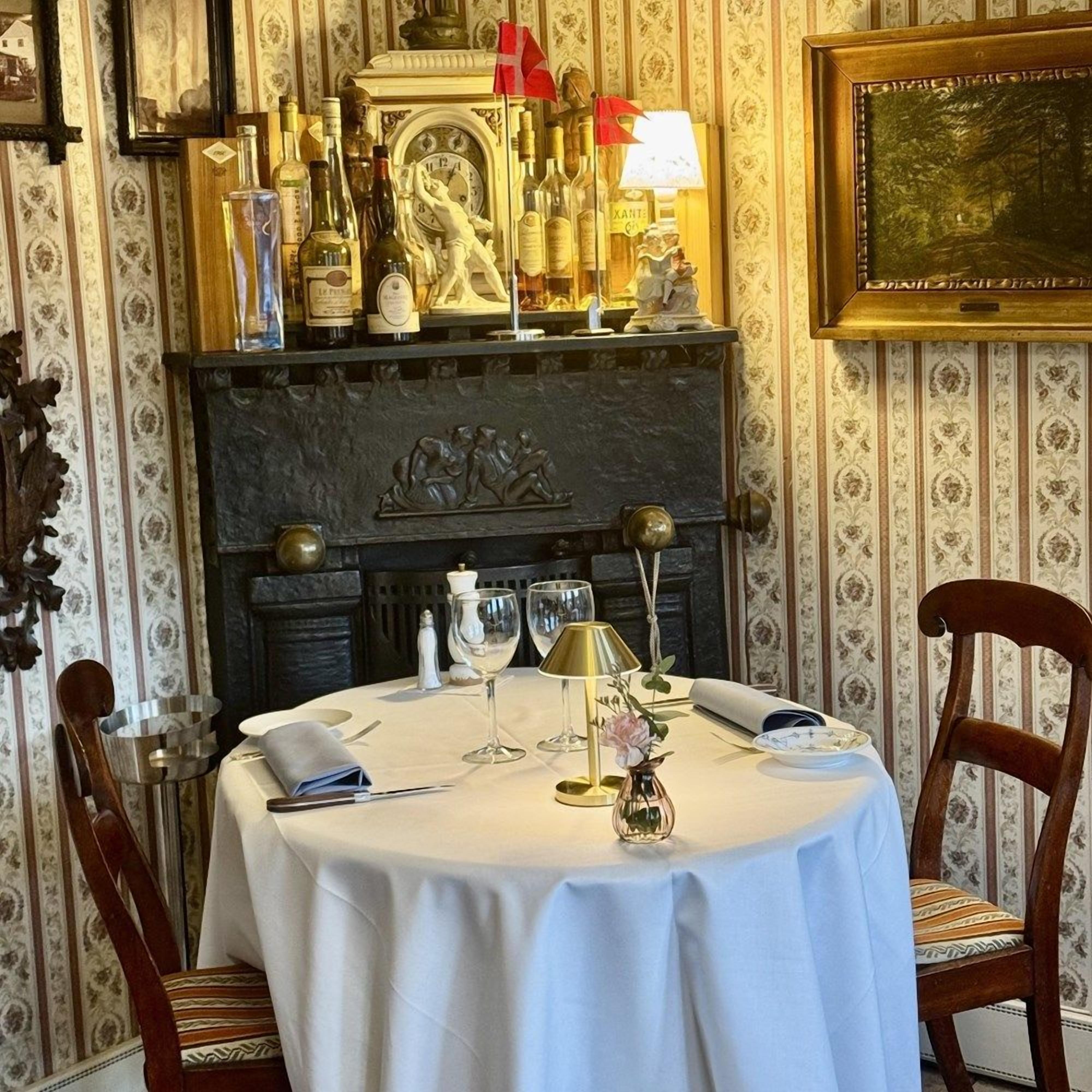 Et spisebord dækket med hvid dug, vinglas, en vase med blomster og en lampe.