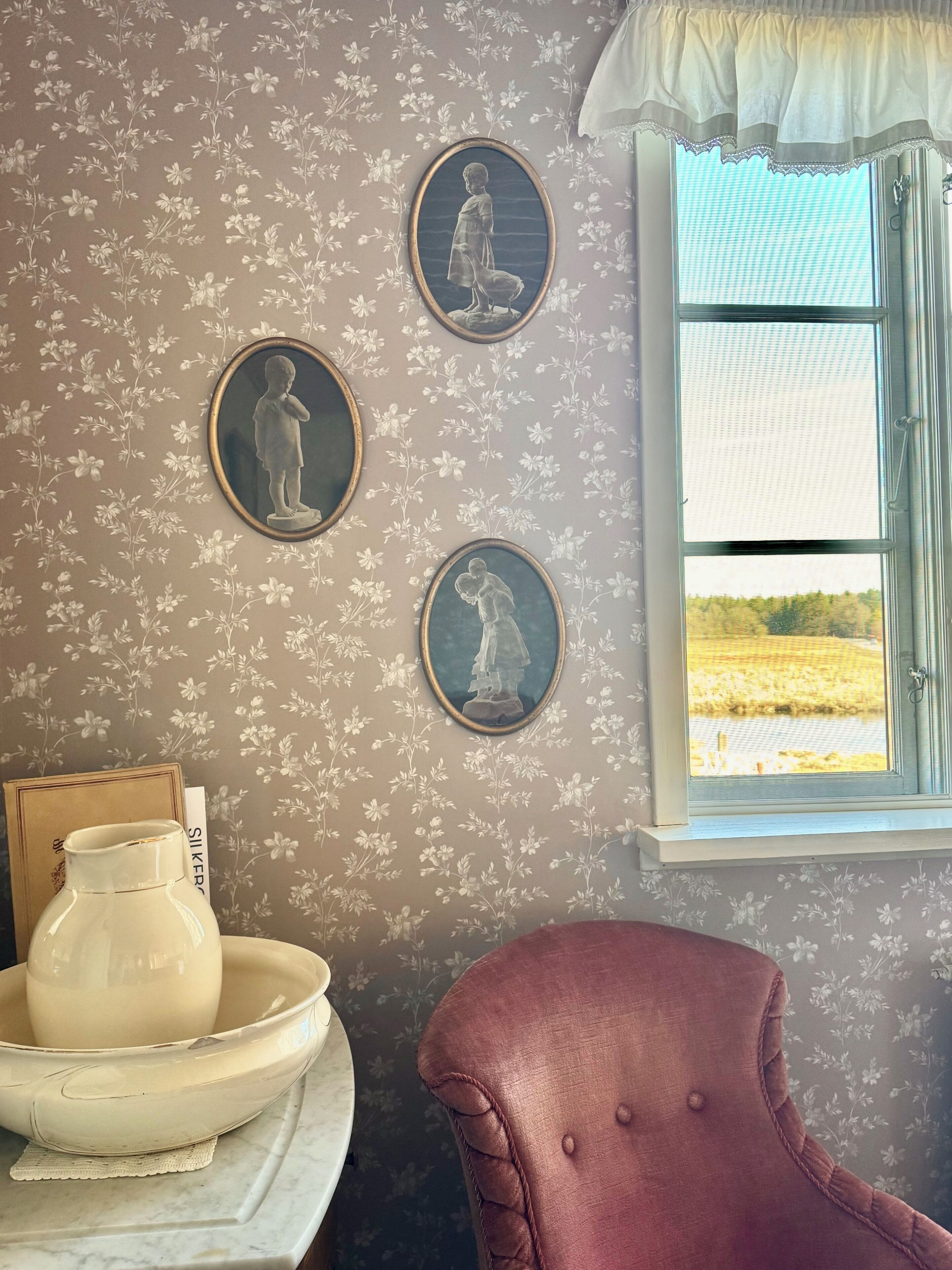 A room with floral wallpaper, a window with a view, a vase, and a chair.