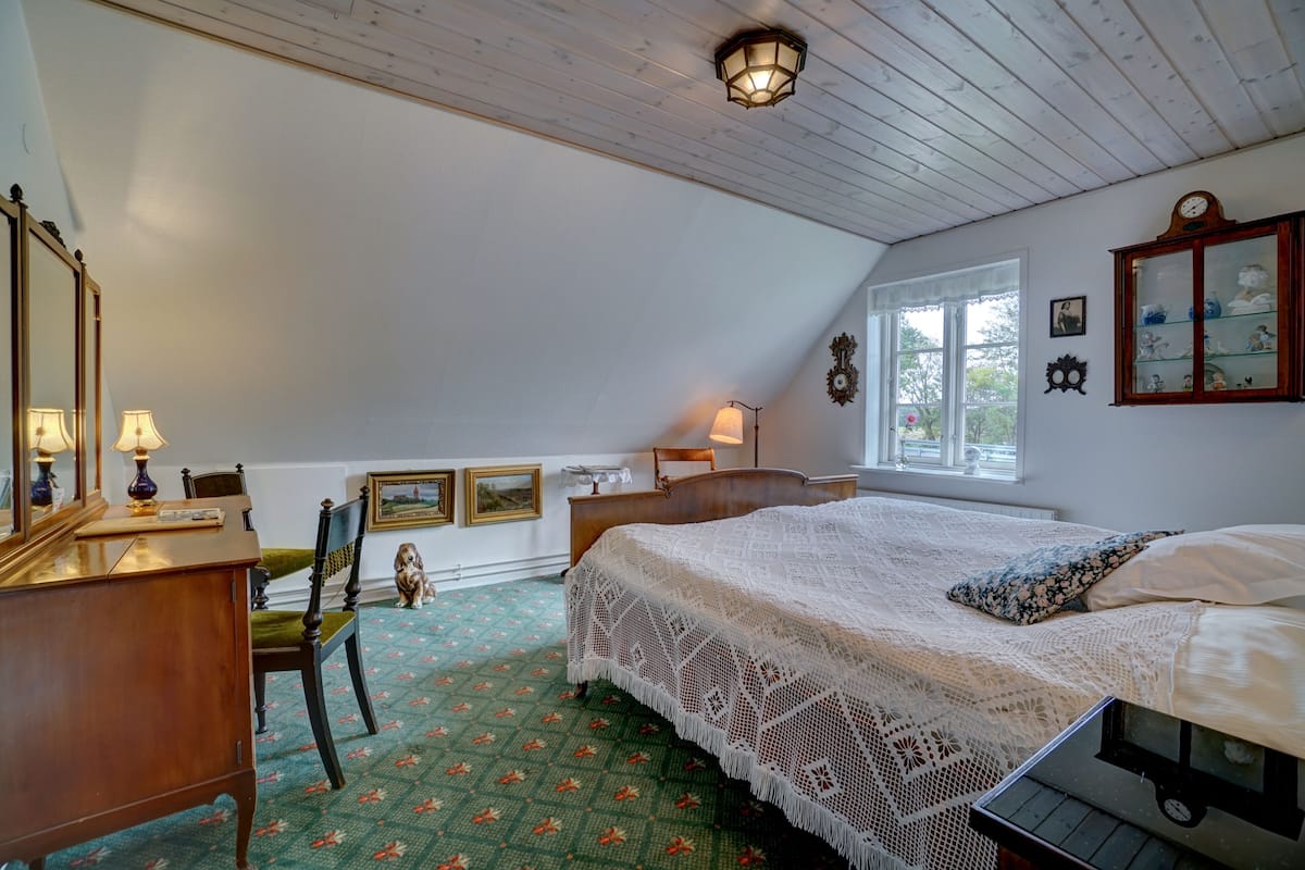 A bedroom with a large bed, a wooden desk, a chair, a dog, and a window with a view of trees.
