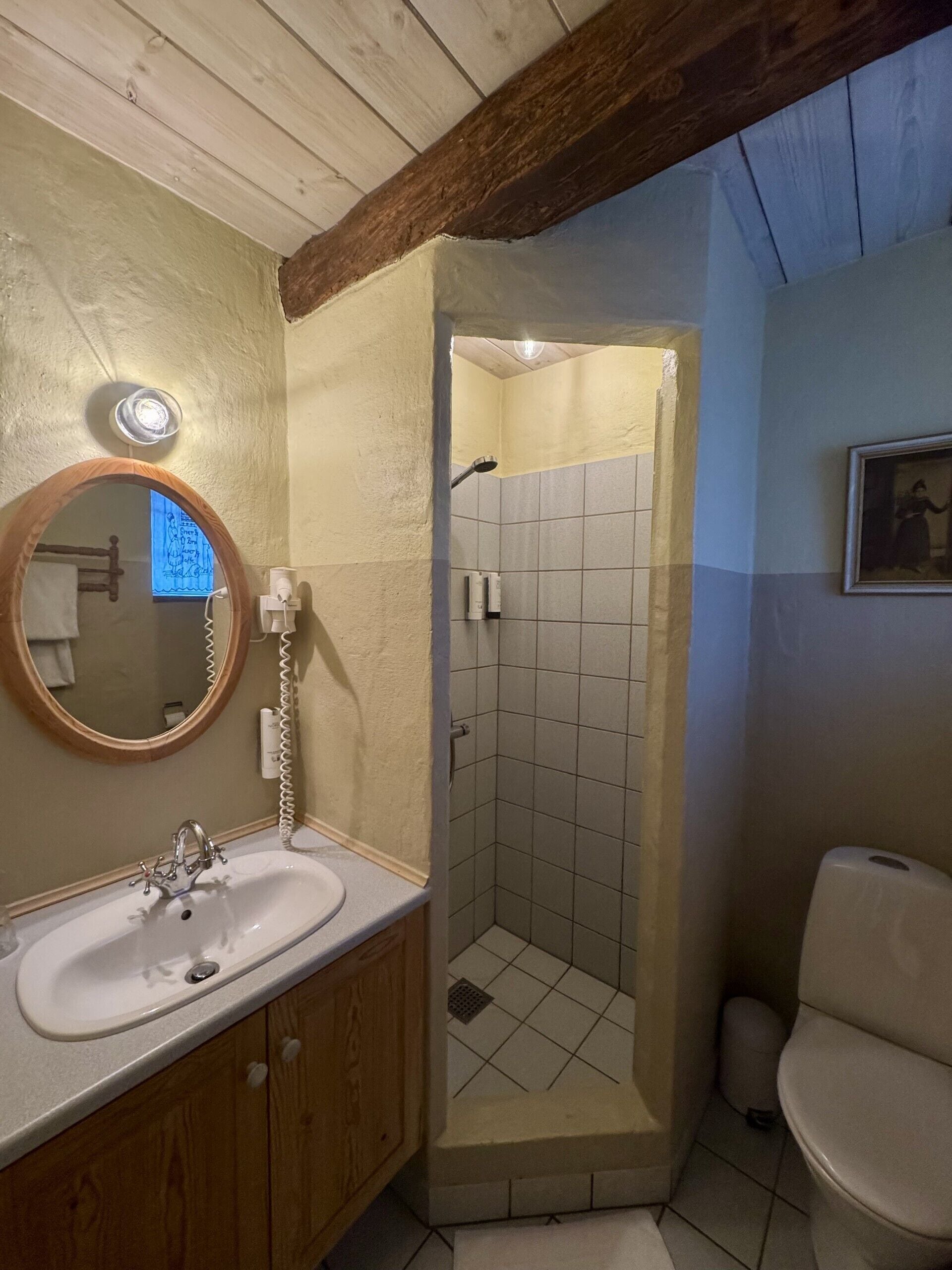 Een badkamer met een houten wastafelmeubel, een ronde spiegel, een douche met betegelde wanden en een toilet.