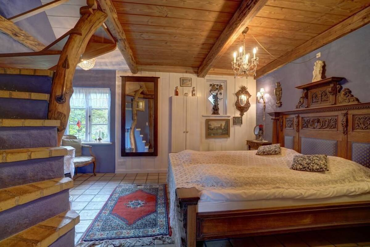 A bedroom with a wooden staircase, a bed with a lace cover, a chandelier, and a fireplace.