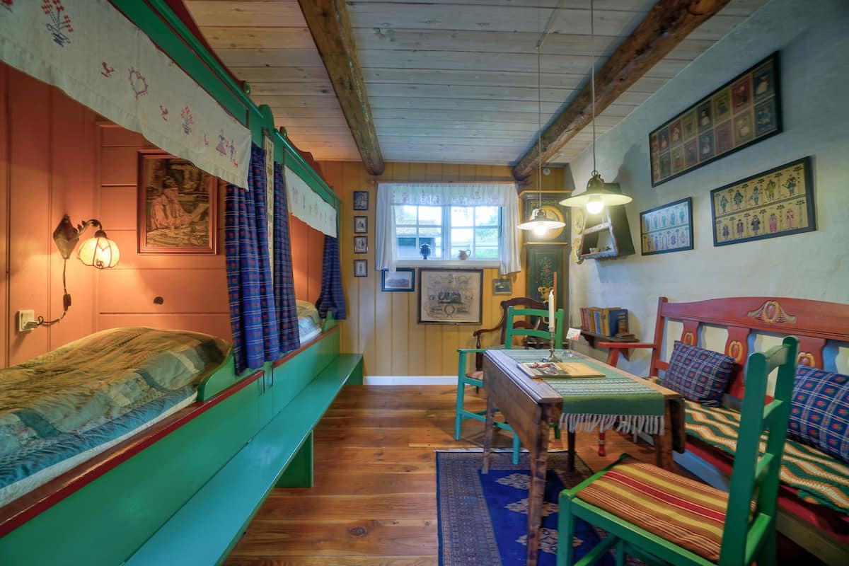 A cozy room with wooden floors, a green bed, a wooden table with chairs, and a window with curtains.