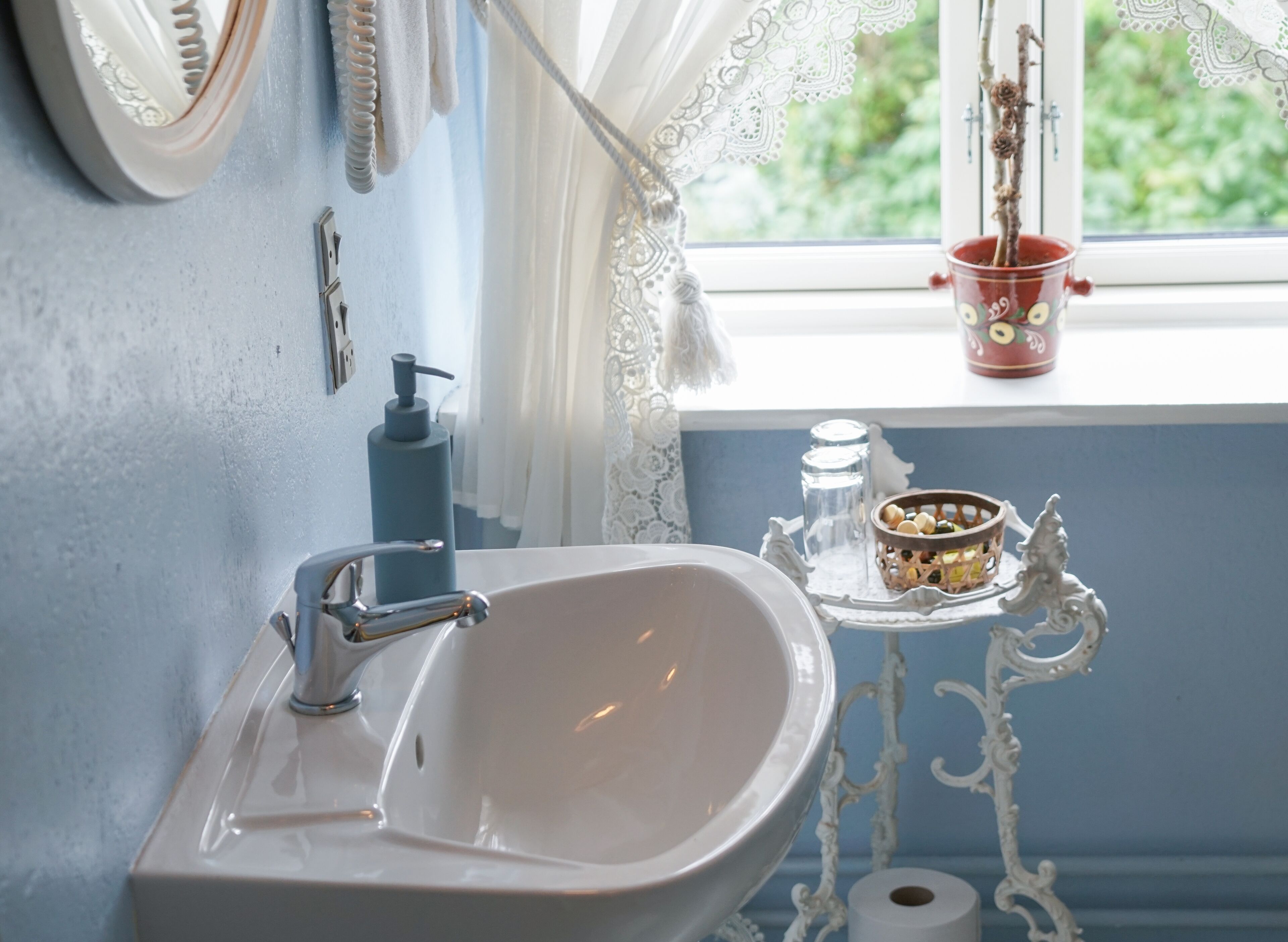 Un baño con un lavamanos blanco, un espejo, una ventana con cortinas de encaje, una maceta decorativa roja y un estante con objetos.