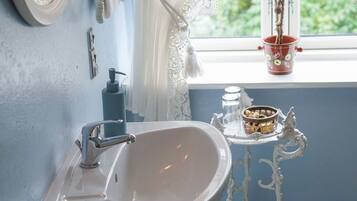 Un baño con un lavamanos blanco, un espejo, una ventana con cortinas de encaje, una maceta decorativa roja y un estante con objetos.