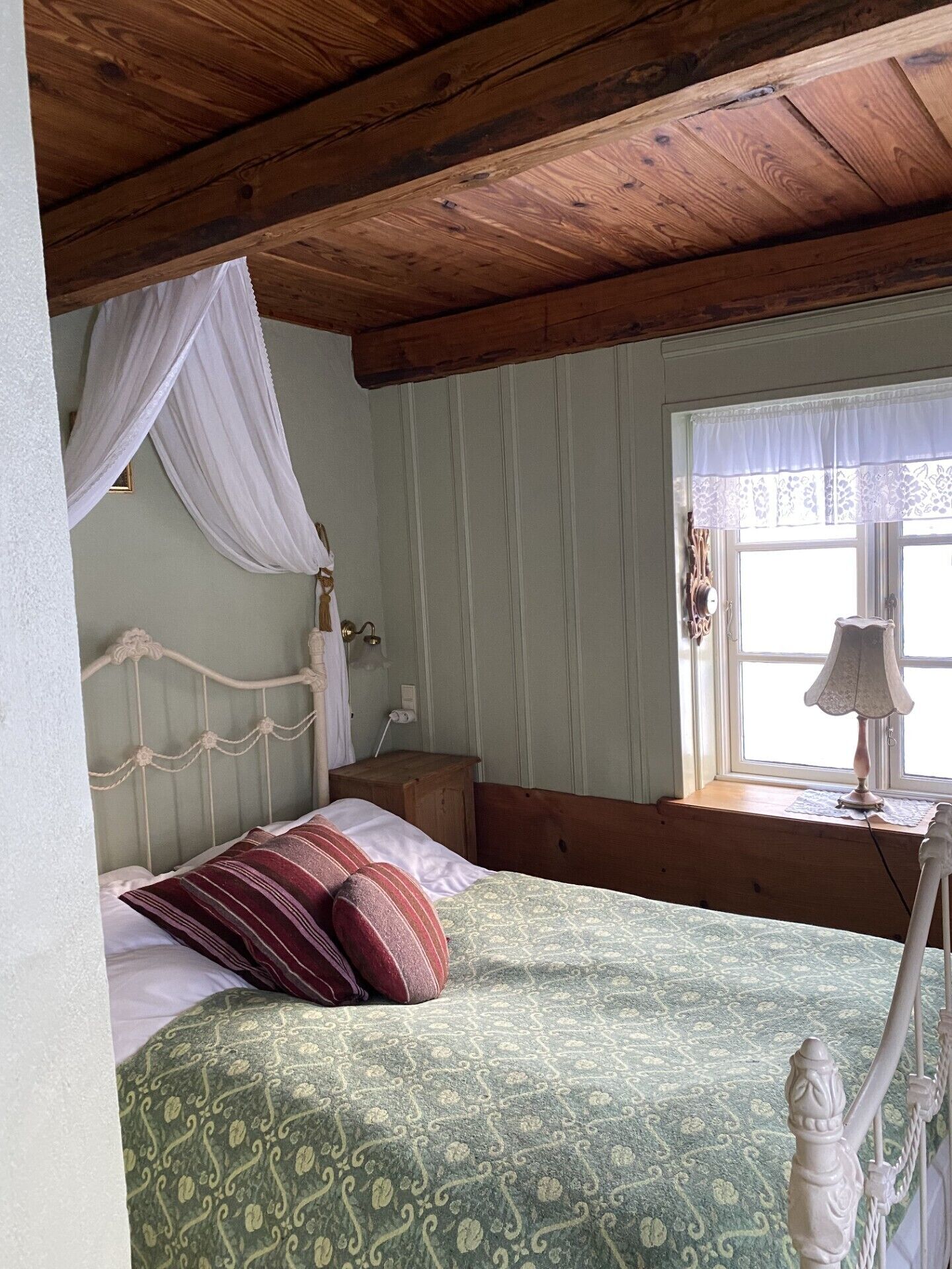 A bedroom with a wooden ceiling, a bed with patterned bedding, a nightstand, a lamp, and a window with a curtain.