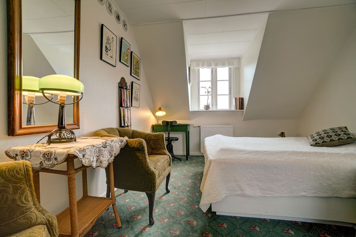 A bedroom with a bed, a chair, a table with a lamp, a mirror, and a window with a view of the outside.