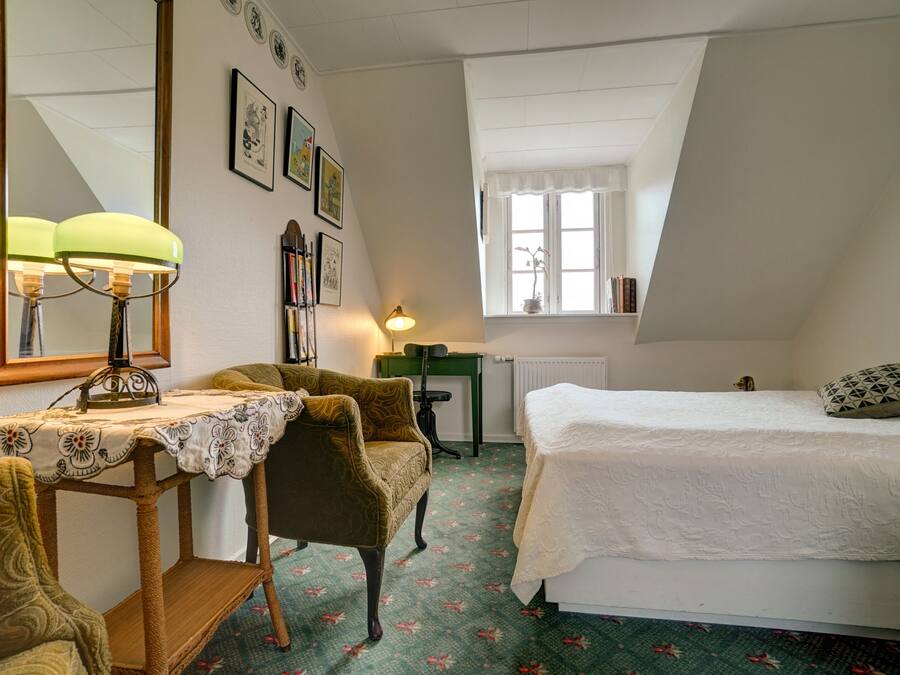 A bedroom with a bed, a chair, a table with a lamp, a mirror, and a window with a view of the outside.