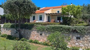 A two-story house with a stone wall in the foreground, a tiled roof, and a garden with various plants and a tree. - Villa Mirothea (Alonissos)