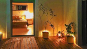 A wooden-floored hallway leading to a bedroom with a bed and a nightstand. There are potted plants and a wall-mounted shelf with books. - Villa Mirothea (Alonissos)