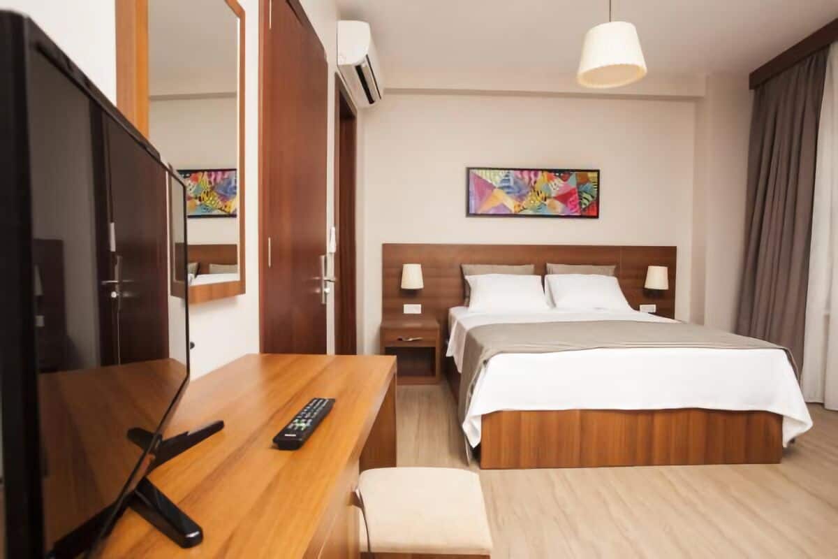 A hotel room with a wooden bed, a desk with a remote control, a bedside table, and a painting on the wall.