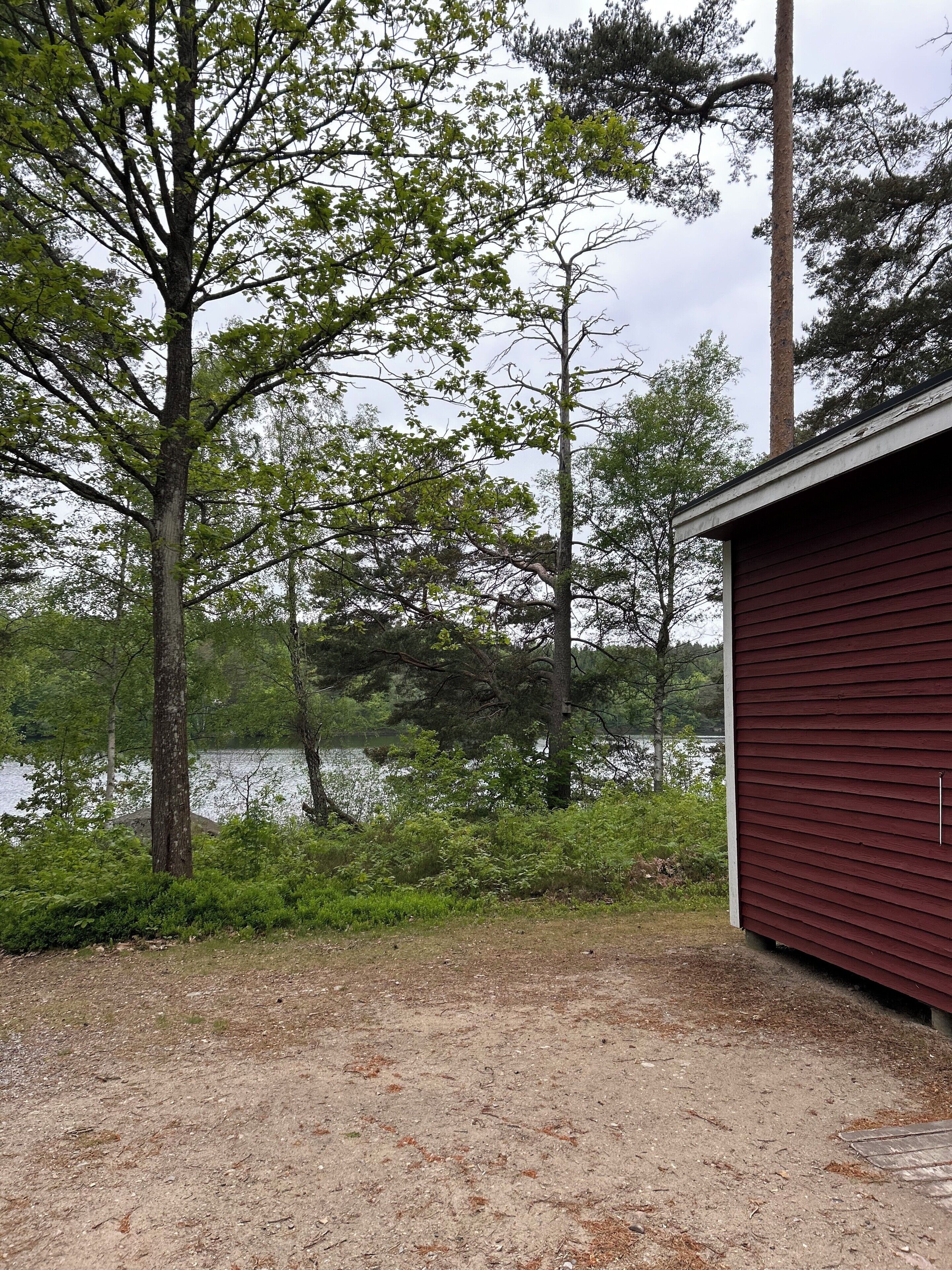 A red building with a dirt ground in front, surrounded by trees and a body of water.