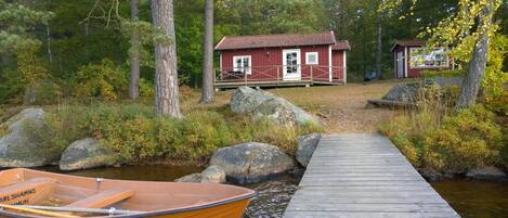 A wooden dock leading to a red cabin with a white door, surrounded by trees and rocks.