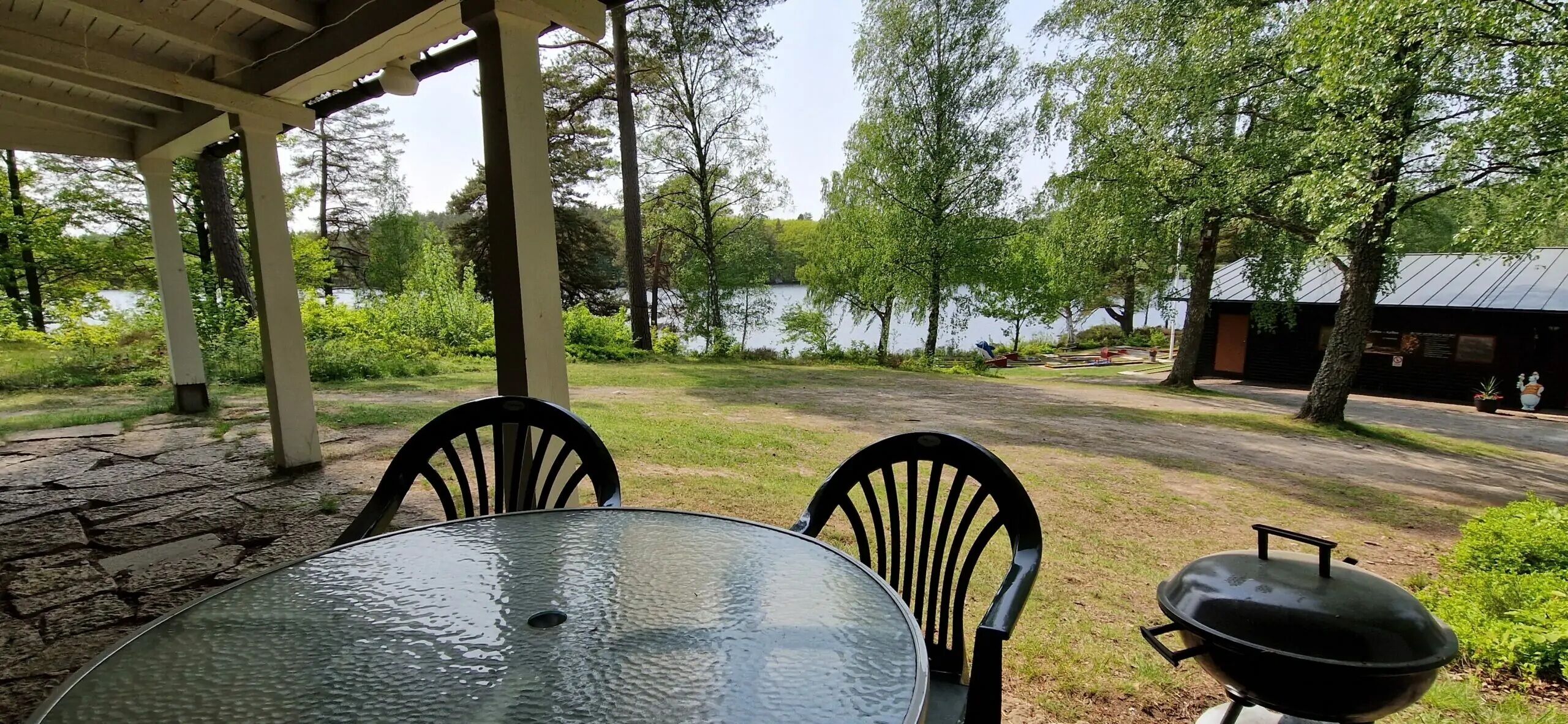 Uma área externa coberta com mesa, cadeiras, churrasqueira e vista para um lago.