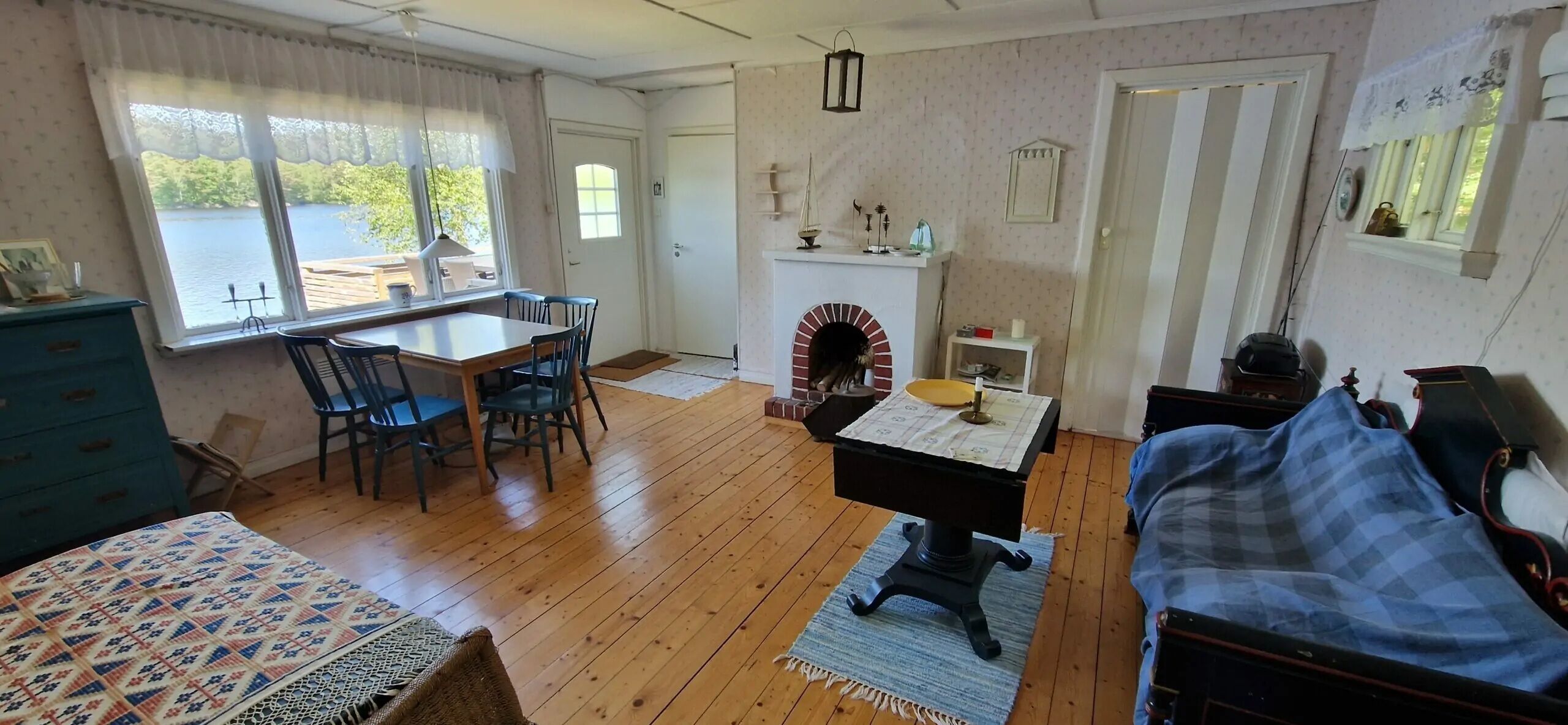 A room with a wooden floor, a dining table with chairs, a fireplace, a bed, and a window with a view of a body of water.