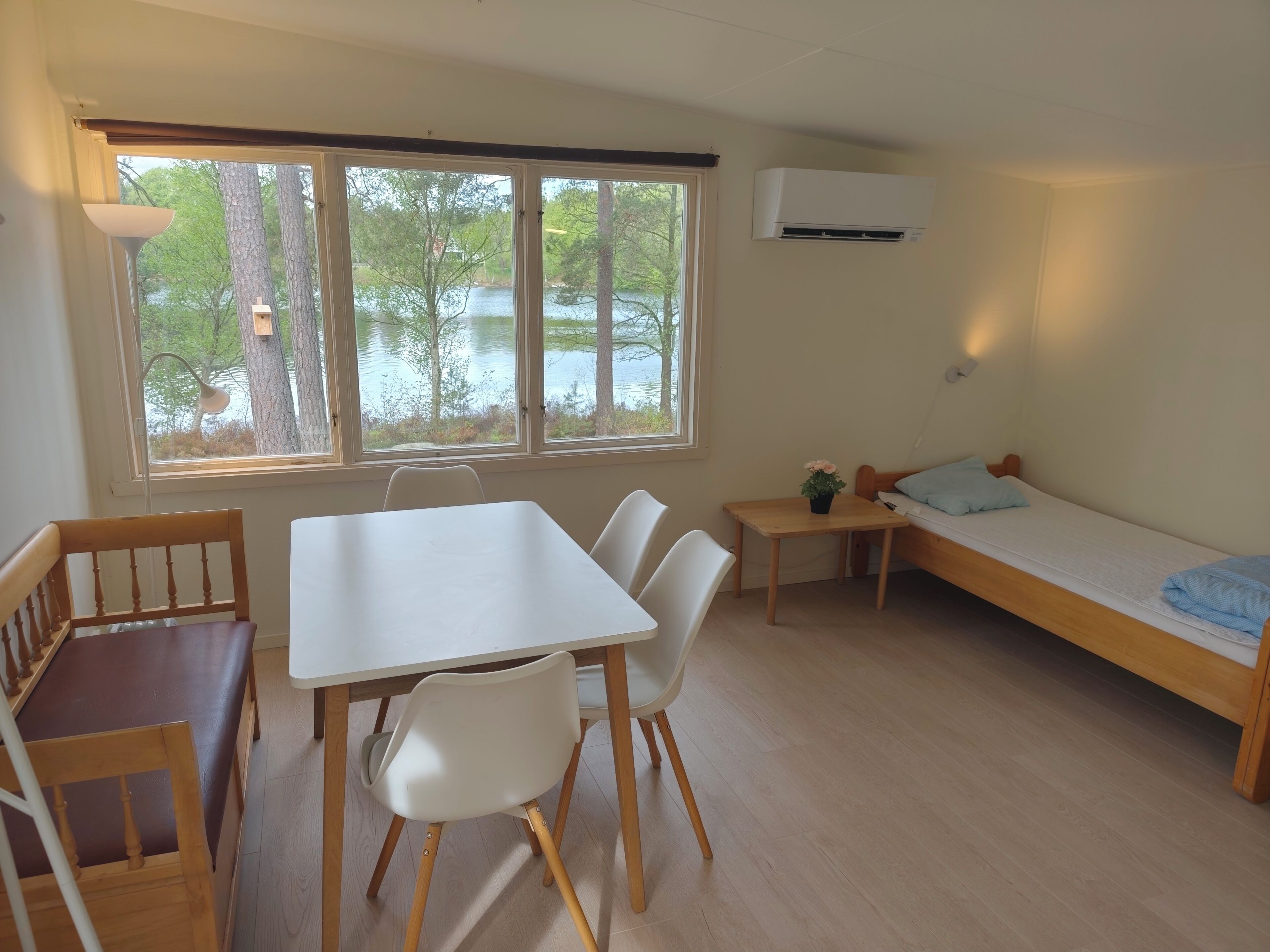A room with a table, chairs, a bed, a window with a view of trees and water, and an air conditioning unit.