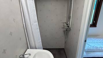 Un baño pequeño con lavamanos, ducha y una cama visible en la habitación contigua.