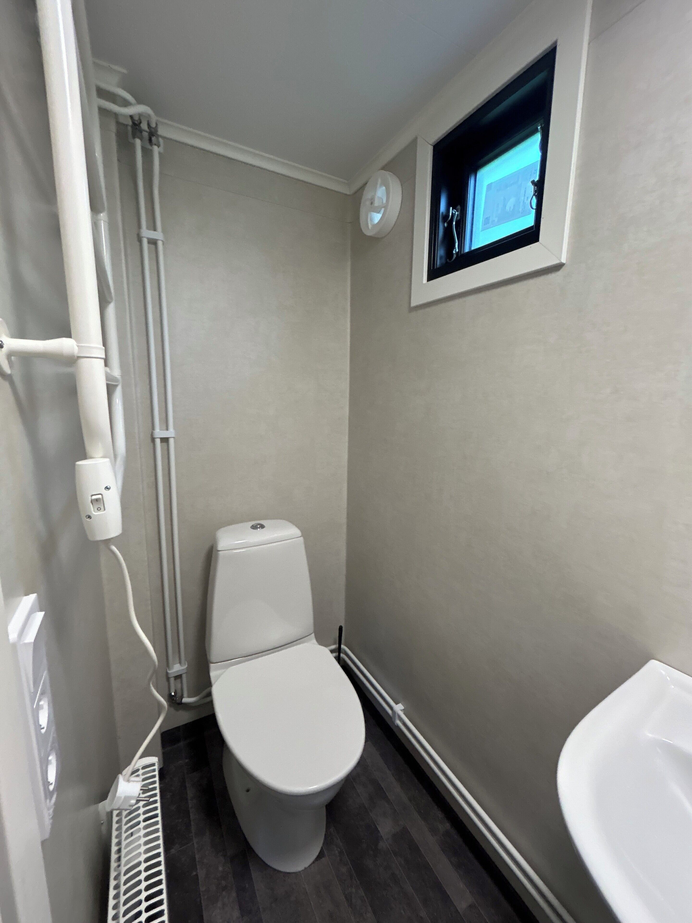 Un baño pequeño con inodoro, lavamanos blanco, televisor montado en la pared y ventilación.