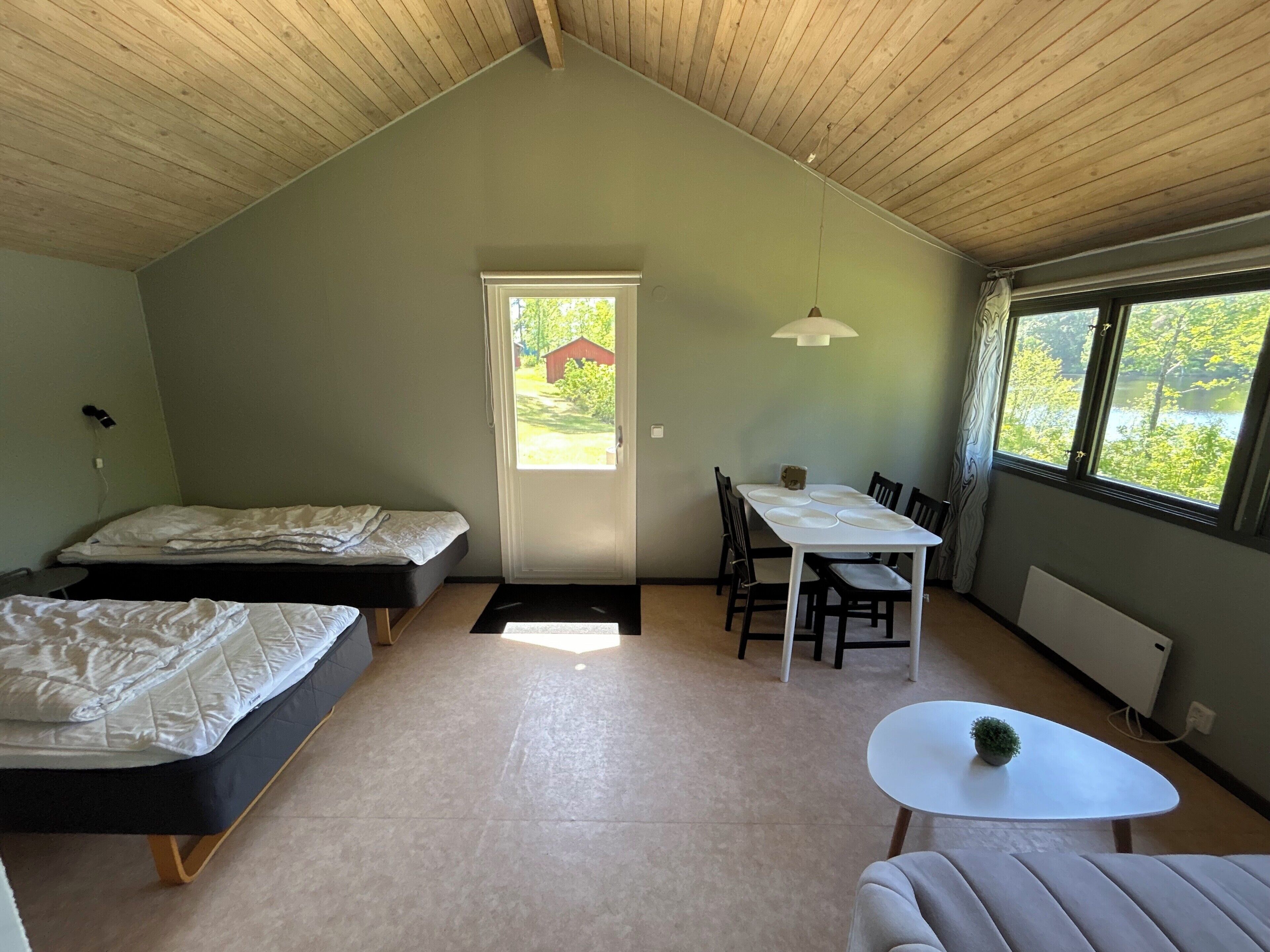 A room with a loft bed, a dining table with chairs, a small white table, and a window with a view of greenery.