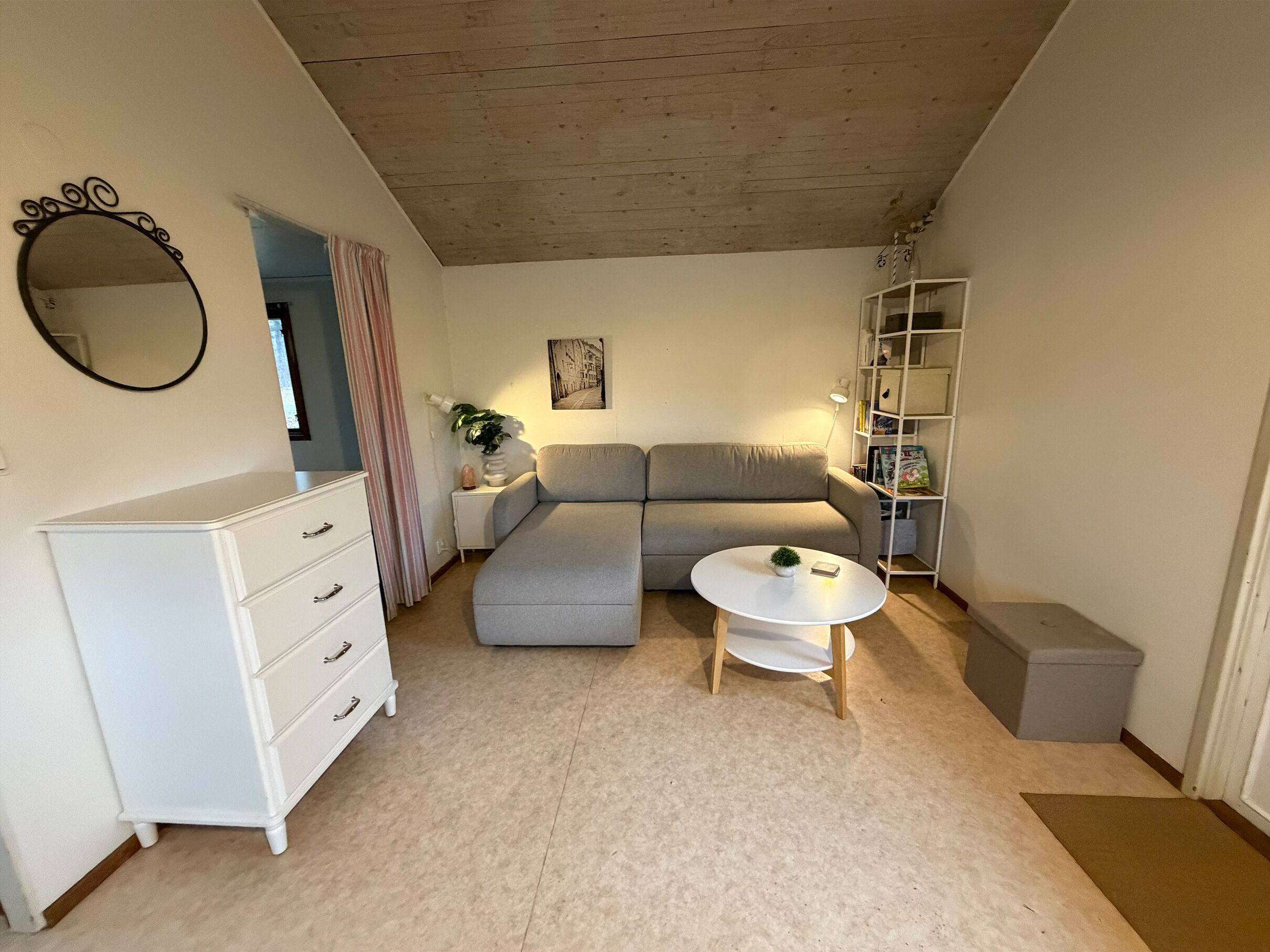 A living room with a sofa, a small round table, a white dresser, a mirror, a bookshelf, and a door.