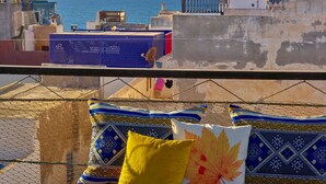 A balcony with a view of the sea, featuring a wooden table with newspapers and a bowl of oranges, and blue and yellow cushions. - Ryad Watier (Essaouira)